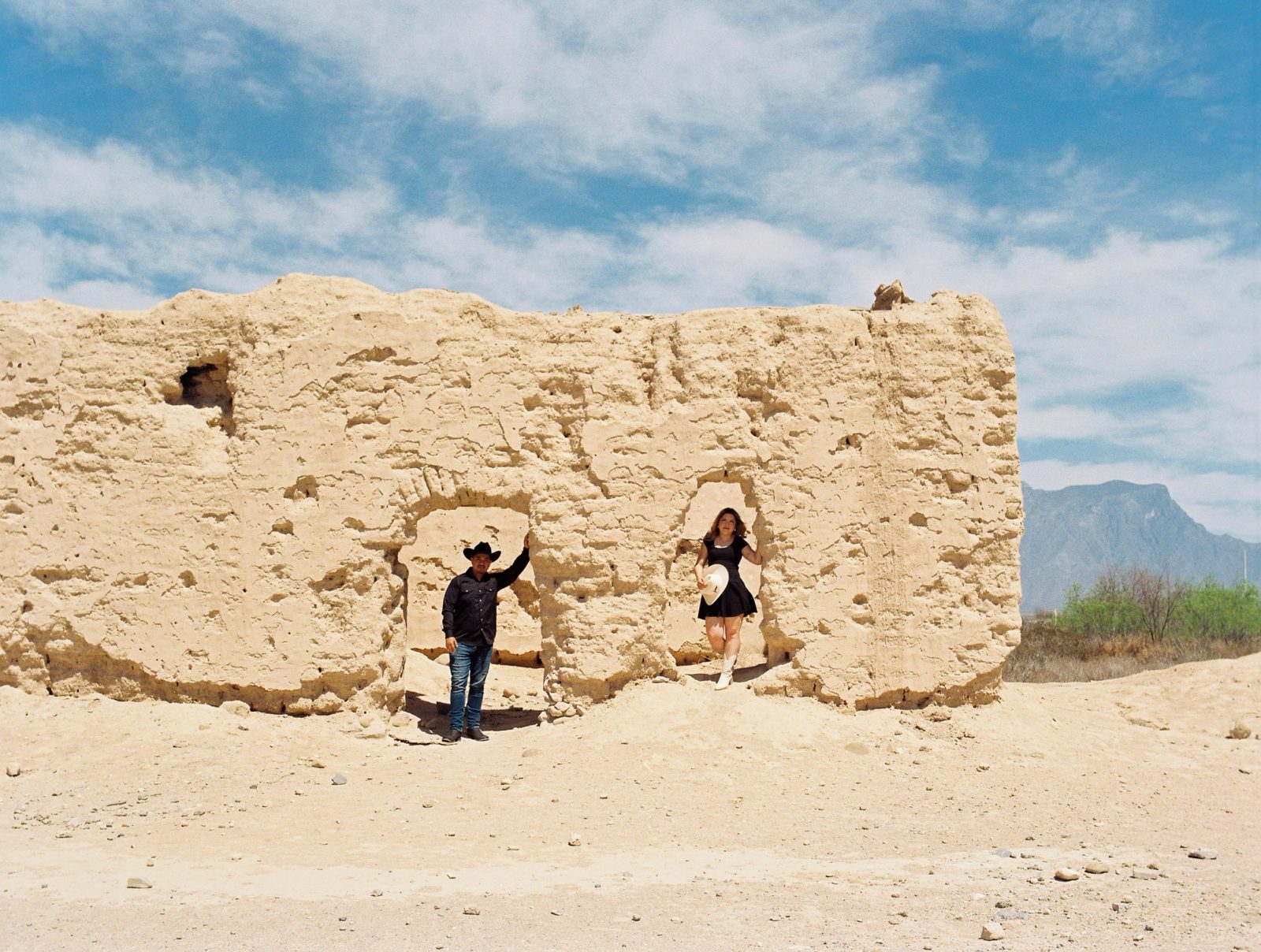 Couple under weathered adobe ruins, dressed in western styling and photographed on film in bright desert light.
