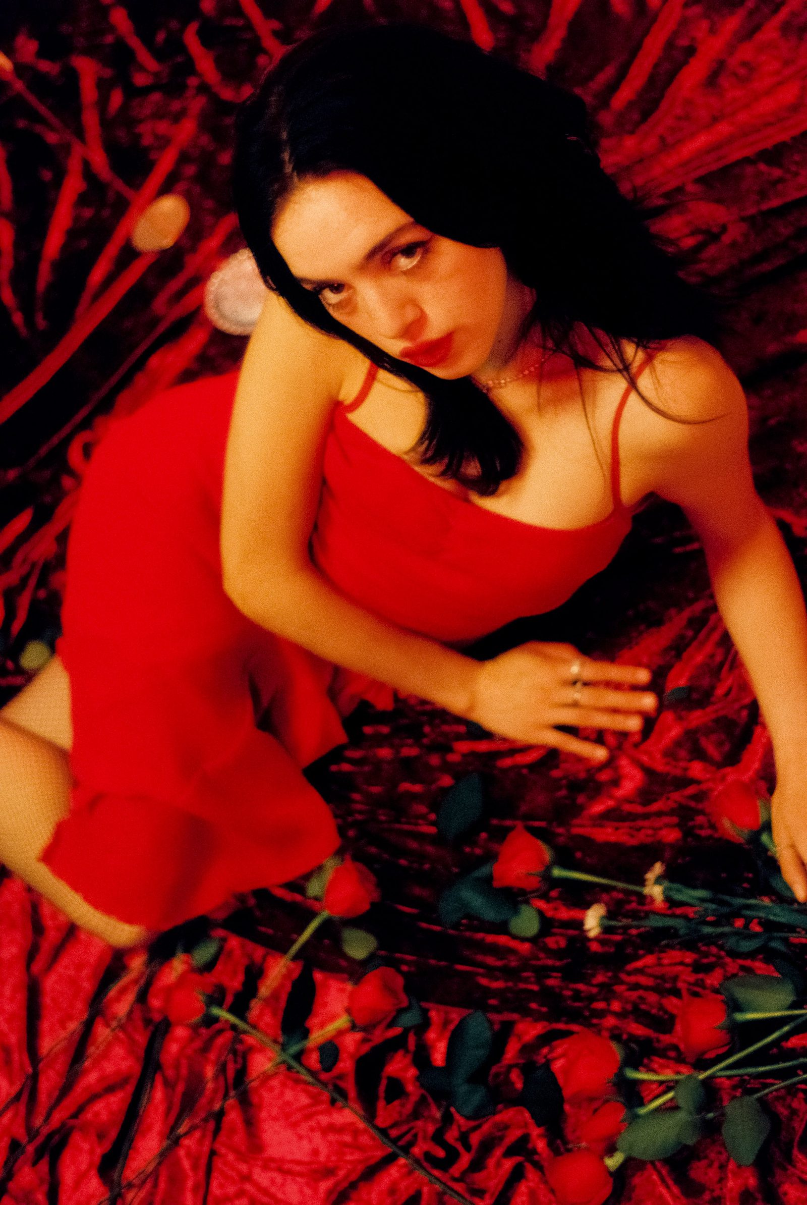Ale Alvarado in a red dress reclining on red velvet surrounded by roses, photographed on film with warm red lighting.