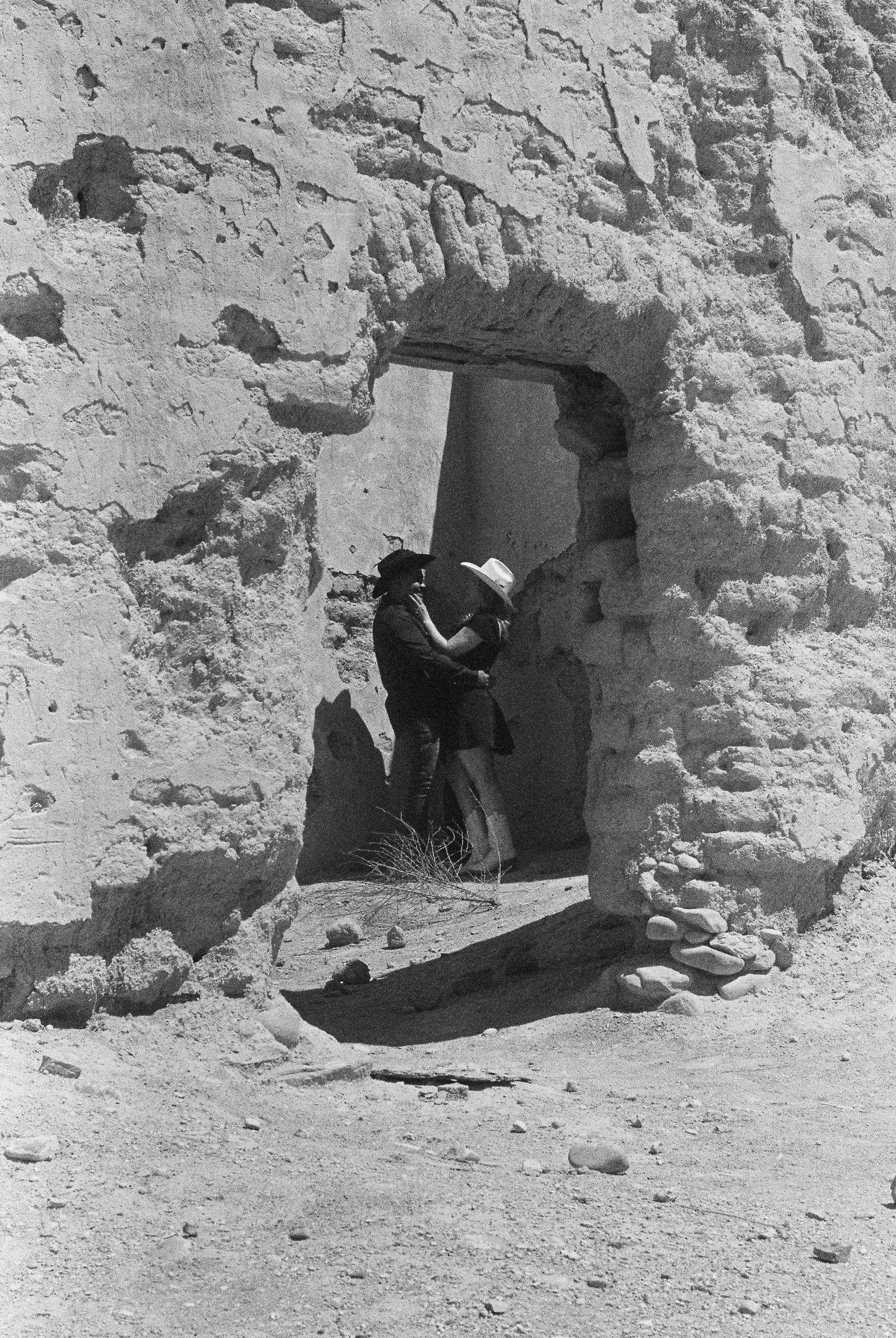 Black and white film portrait of a couple in cowboy hats standing under an adobe ruin doorway in a desert landscape.