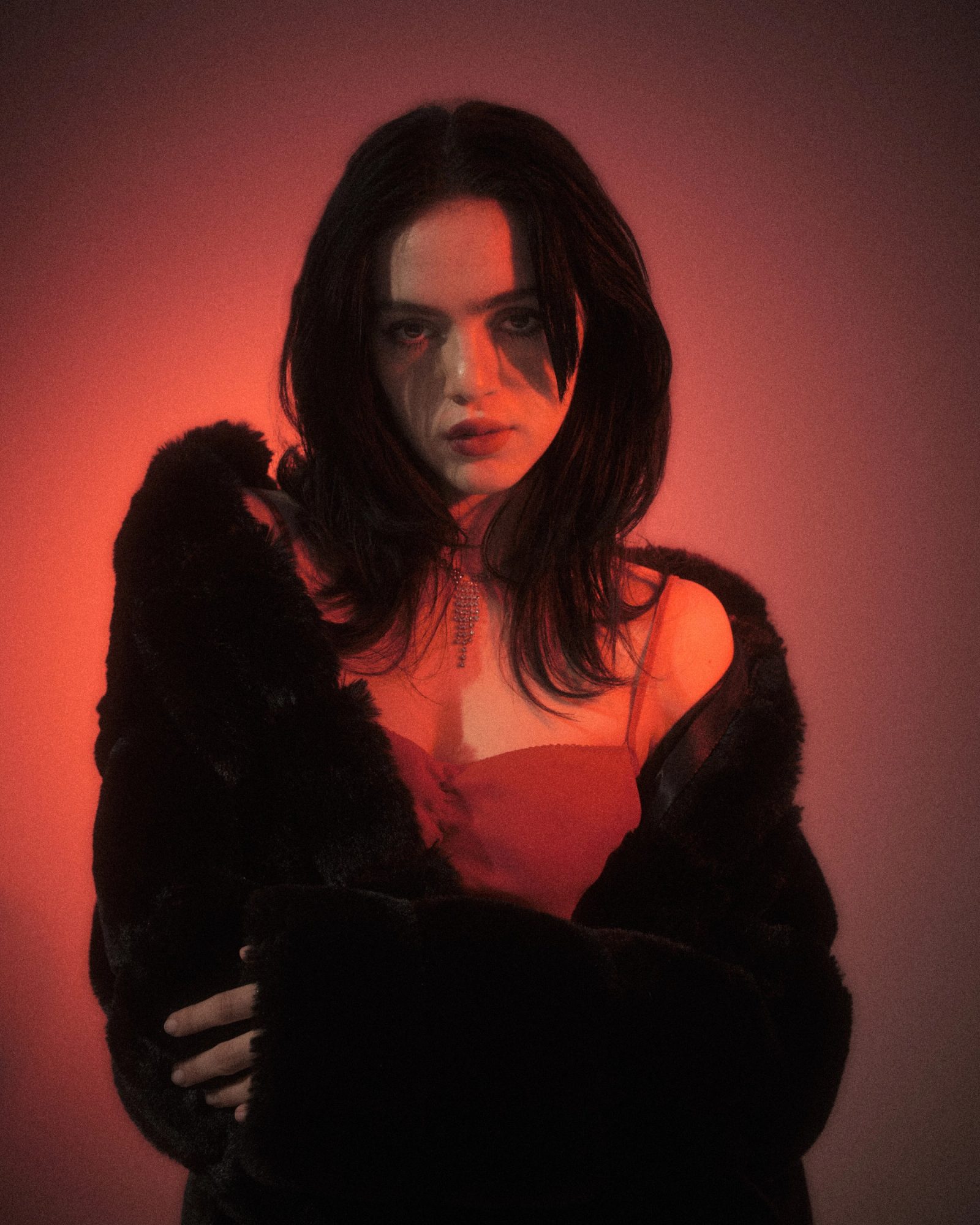 Ale Alvarado posing in a red dress and black fur coat under dramatic red studio lighting.
