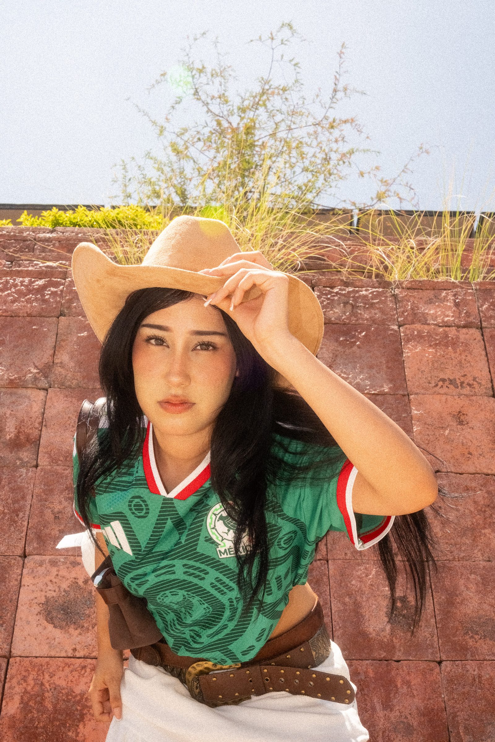 Liz con sombrero y el jersey de la Selección Mexicana en un fondo de losetas de adobe agachada mirando a la cámara