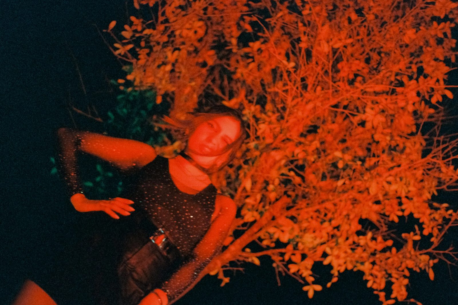 Angela Hernandez leaning beside red-lit foliage at night, photographed in a punk-inspired outfit with a cinematic grainy look.