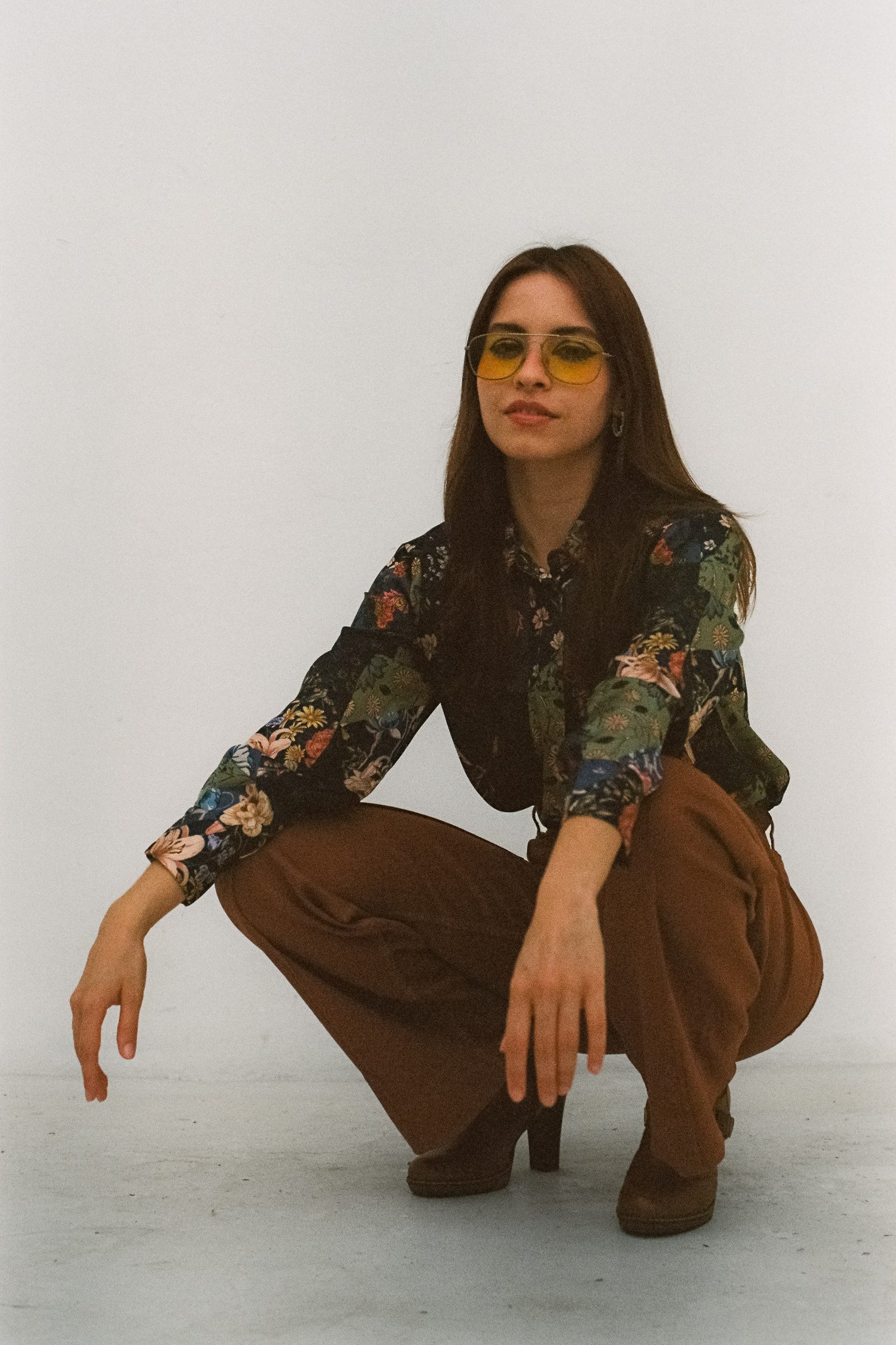 Editorial portrait of Karen Cantu crouching while wearing yellow-tinted sunglasses, a floral shirt, and brown pants, with her arms relaxed forward against a white background.