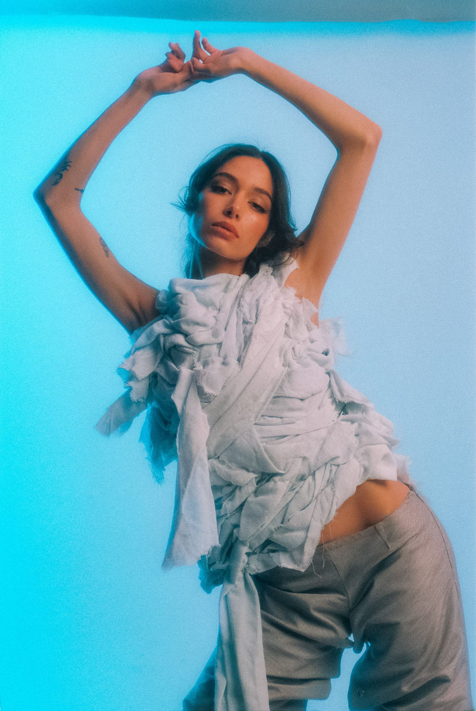 Michelle Malacara standing with arms raised in a sculptural pale fabric top and gray pants, photographed on film with cool blue studio light.