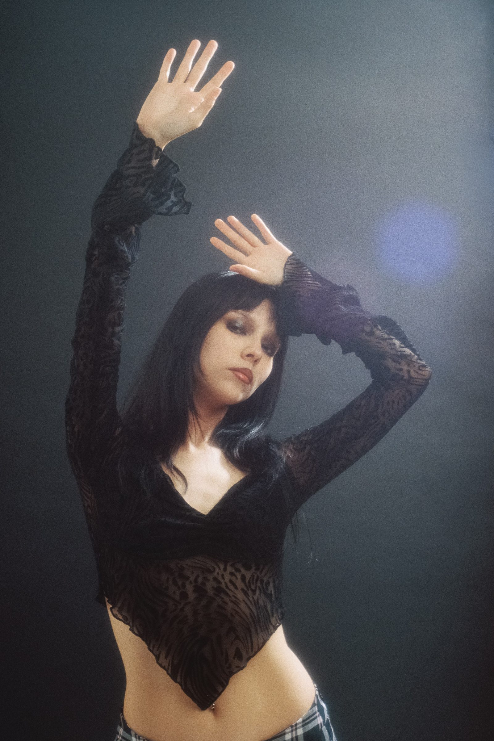 Dany Mass posing with raised hands in a black sheer top, photographed on film with soft light and a dark studio background.