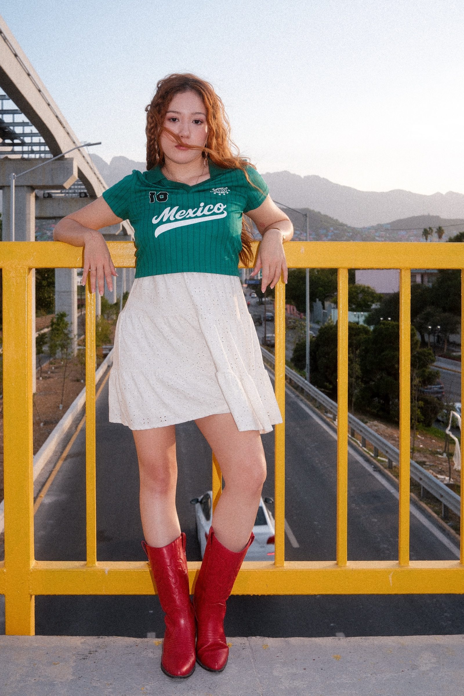 A redhaired girl dressed in Mexico's colors in a yellow bridge with the road under her and the mountains on the back