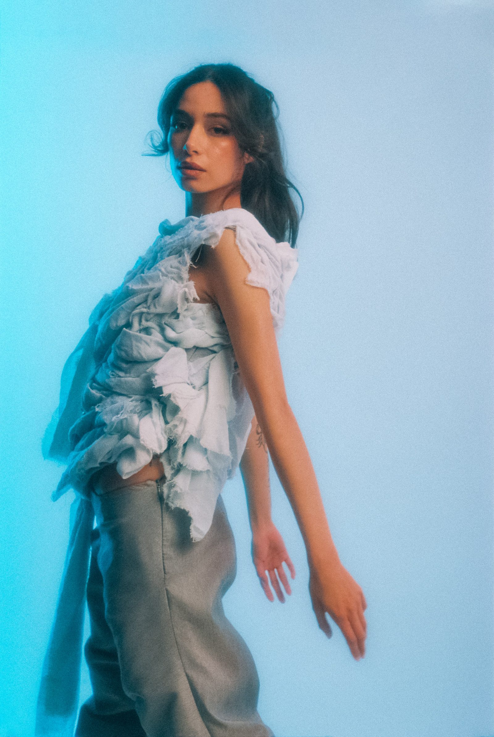 Michelle Malacara in profile wearing a sculptural pale fabric top and gray pants, photographed on film against a soft blue studio backdrop.