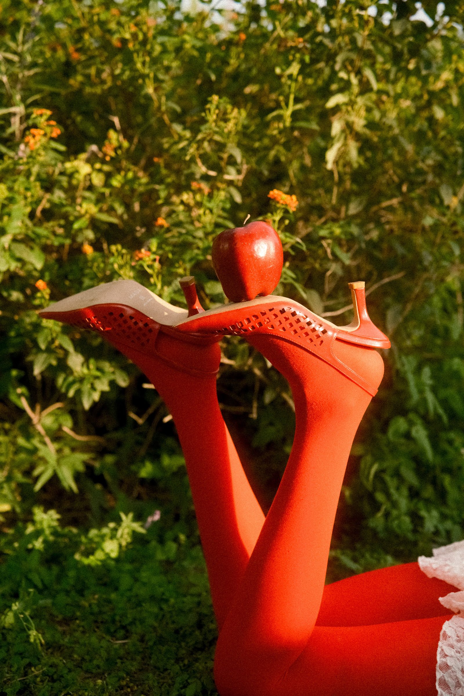 A model raises her legs in red tights while holding a red apple against a green garden background.