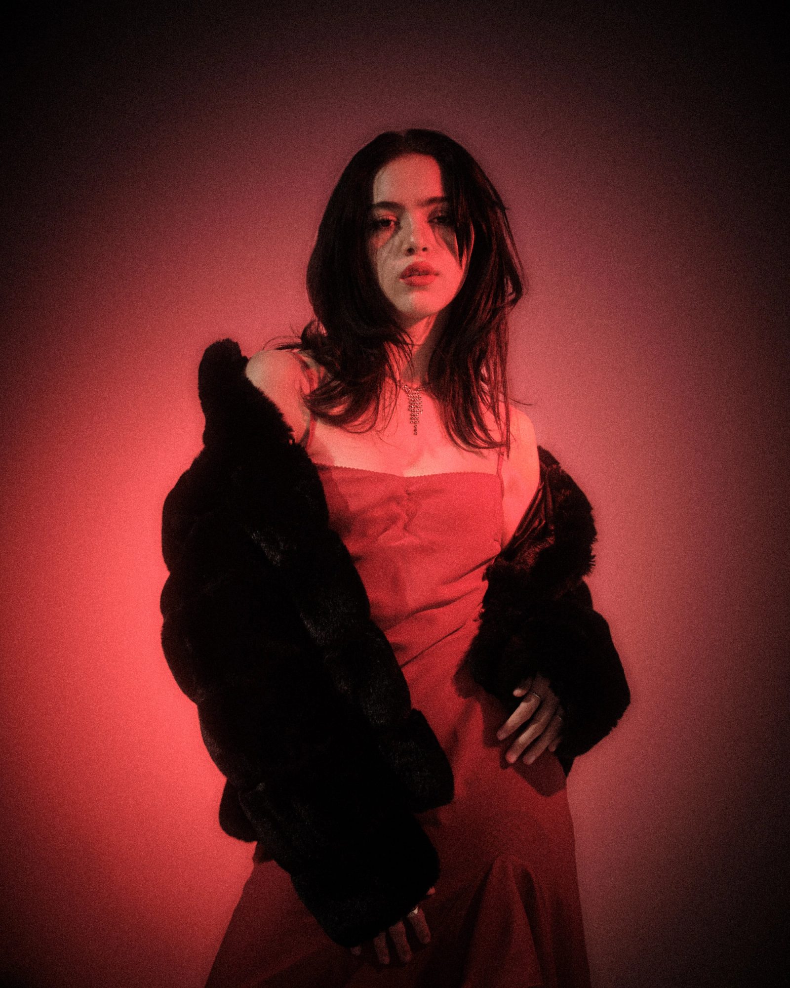 Ale Alvarado standing in a red dress and black fur coat, photographed in saturated red studio light.
