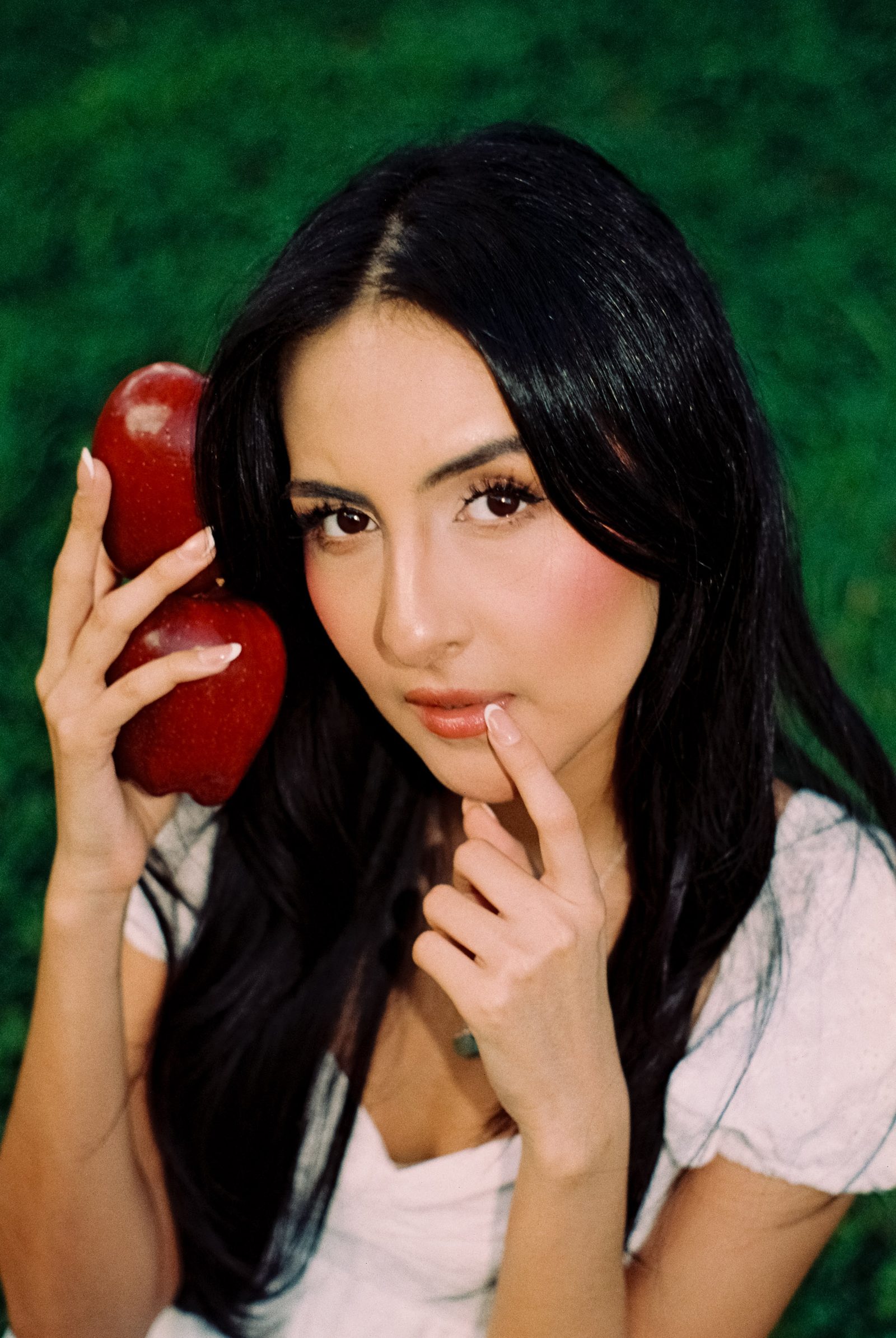 Portrait of a model in a white dress holding a red apple near her face against a green background.