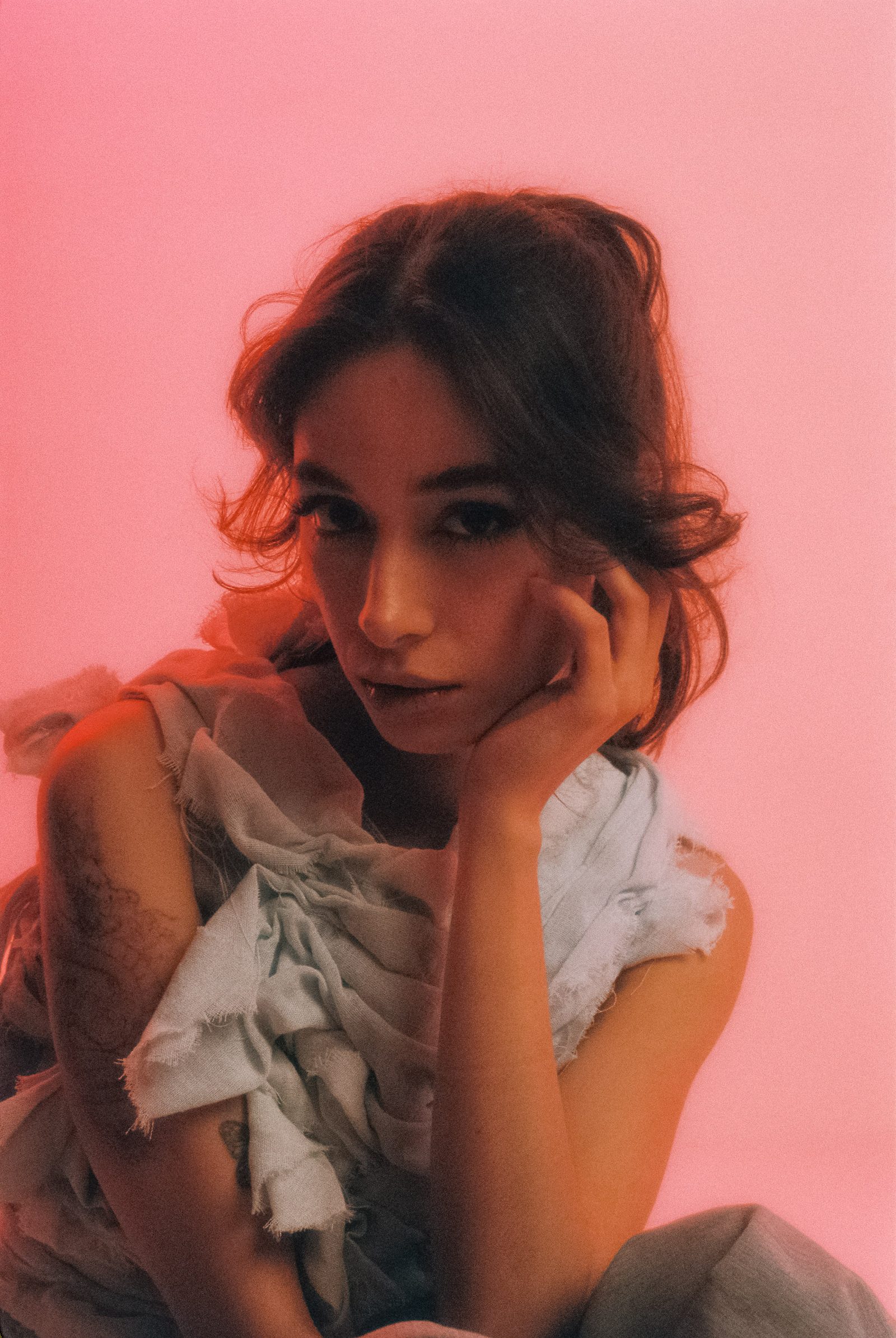 Michelle Malacara resting her face on one hand in a sculptural pale fabric top, photographed on film against a pink studio backdrop.