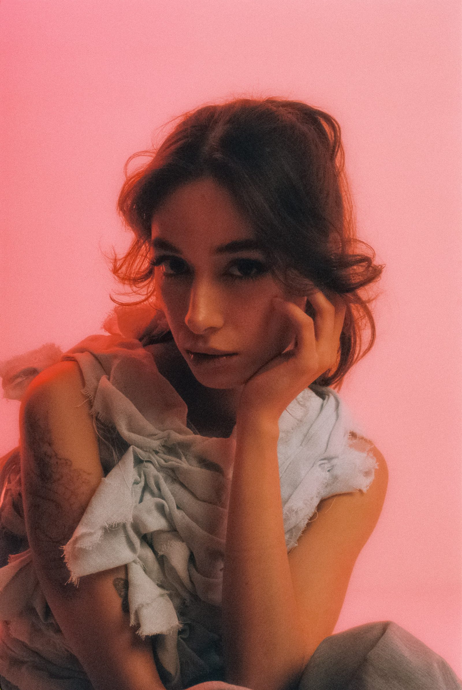 Michelle Malacara resting her face on one hand in a sculptural pale fabric top, photographed on film against a pink studio backdrop.