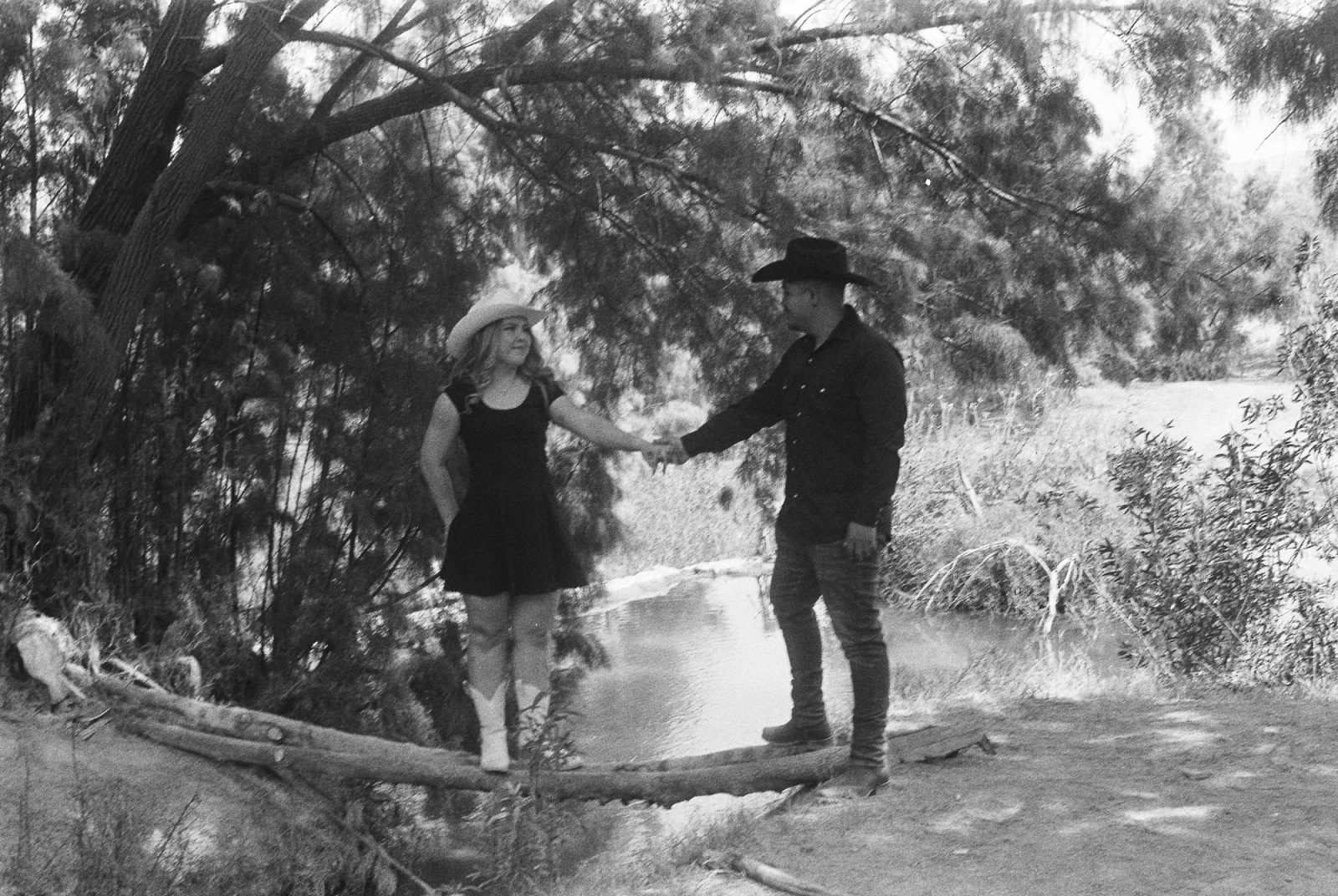 Black and white film portrait of a couple in cowboy hats holding hands beneath trees beside a river.