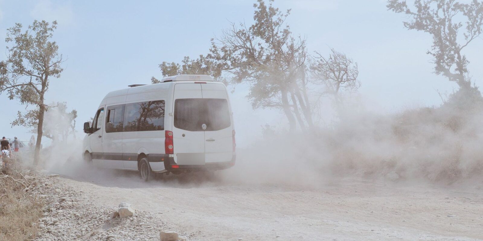White passenger van driving down a dusty gravel road past dry trees