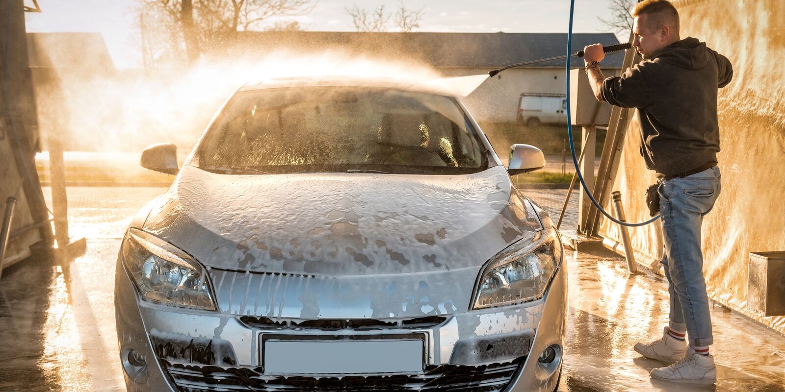 Man pressure washing a foam-covered car in golden light