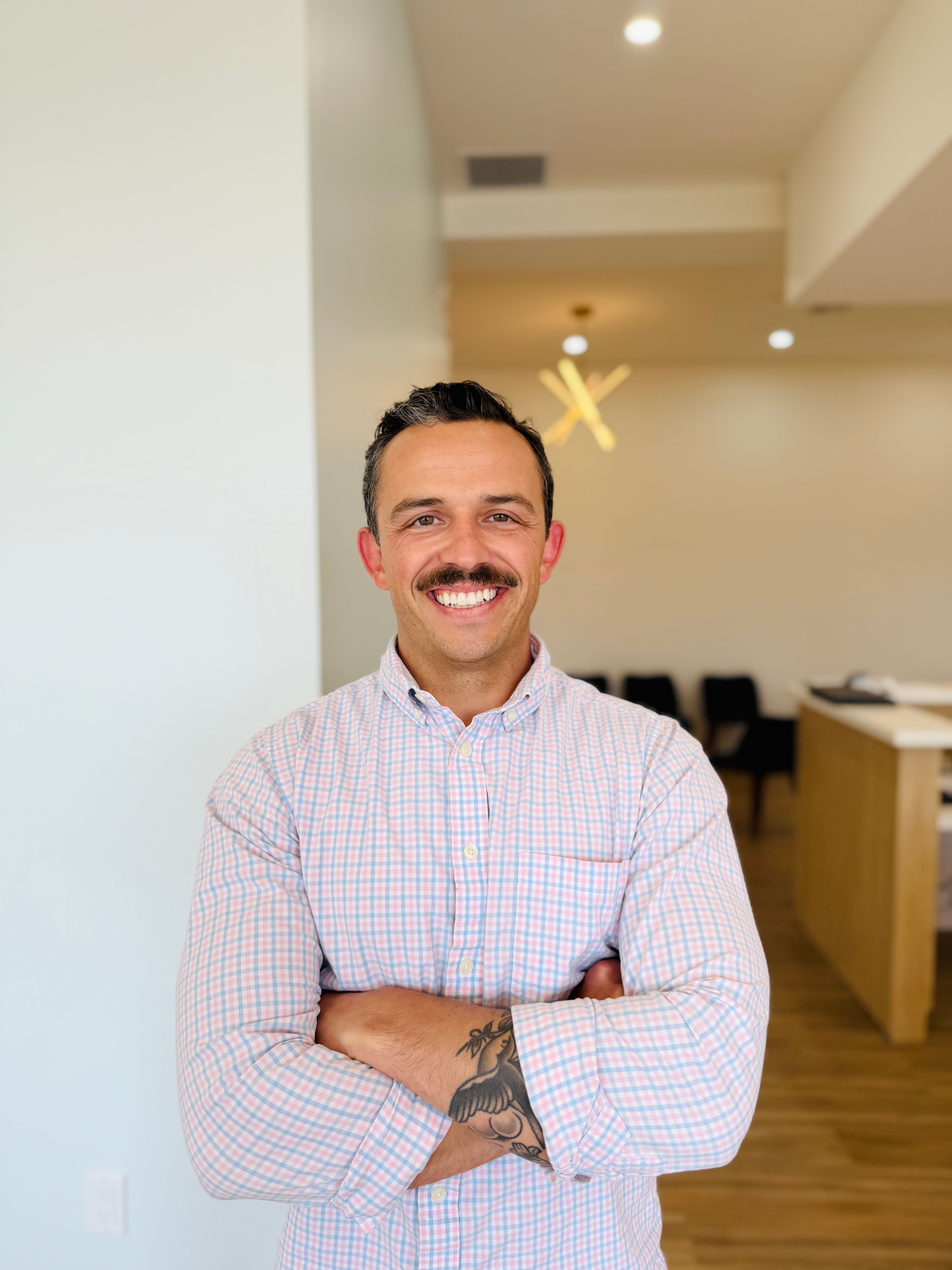 Smiling man with a mustache wearing a light pink and blue checkered button-down shirt, standing indoors with arms crossed in a bright, modern office setting.