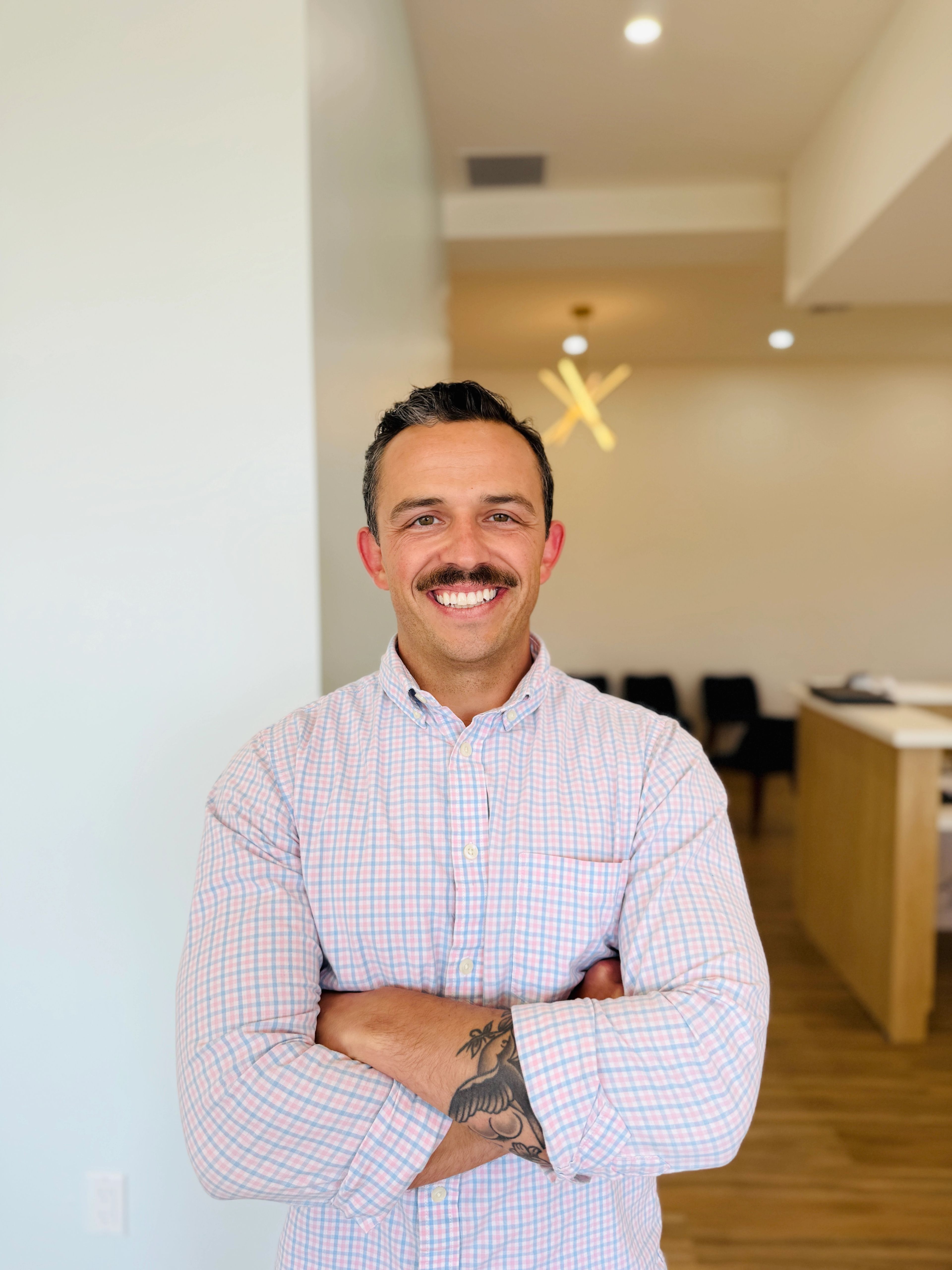 Smiling man with a mustache wearing a light pink and blue checkered button-down shirt, standing indoors with arms crossed in a bright, modern office setting.