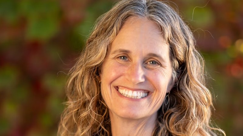 Robin Kaplan smiling outdoors in a navy blouse, with shoulder-length hair and a softly blurred background