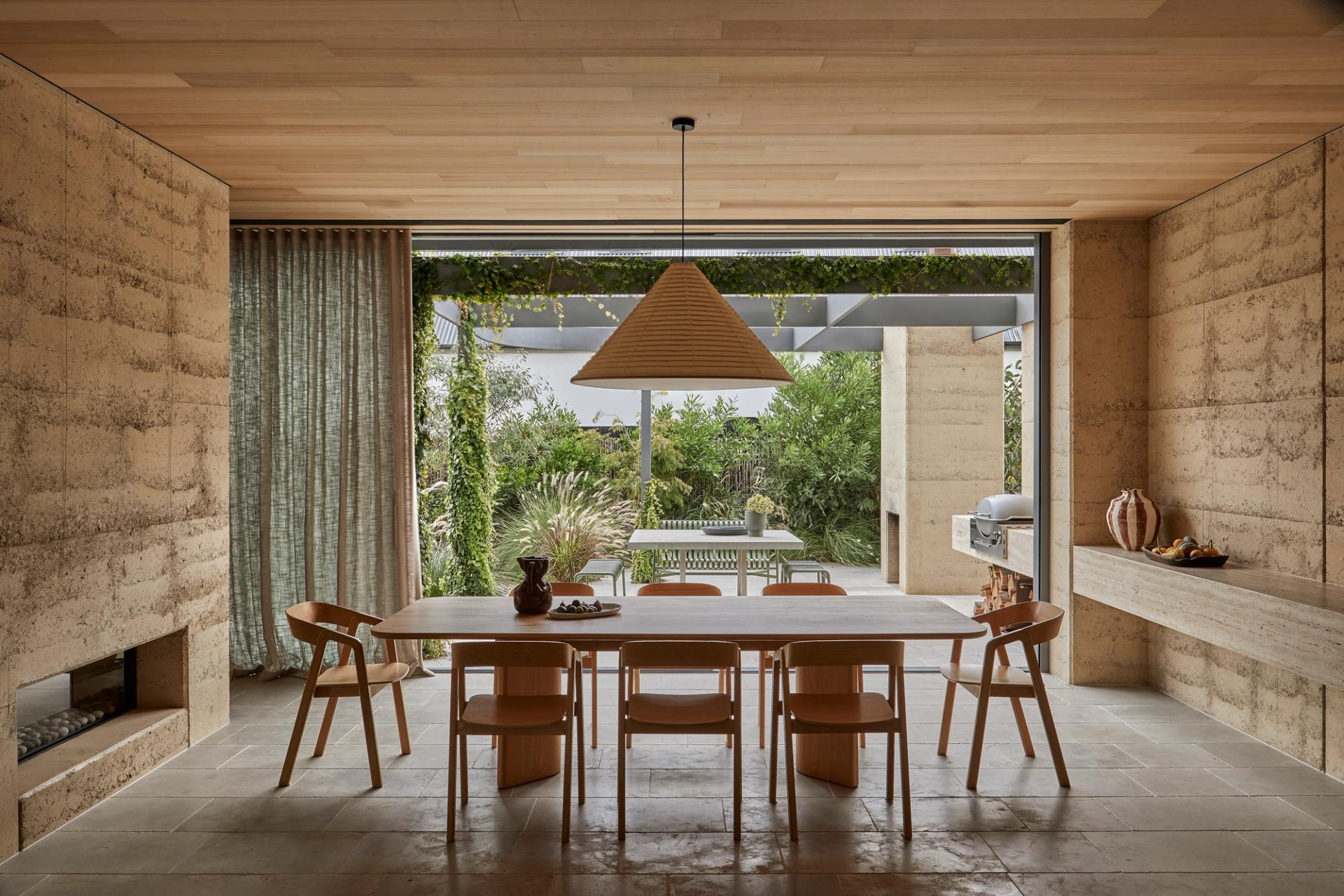 Dining area at Barwon Dunes House opening to sunlit terrace with rammed earth walls, timber lined ceiling and seamless indoor-outdoor living