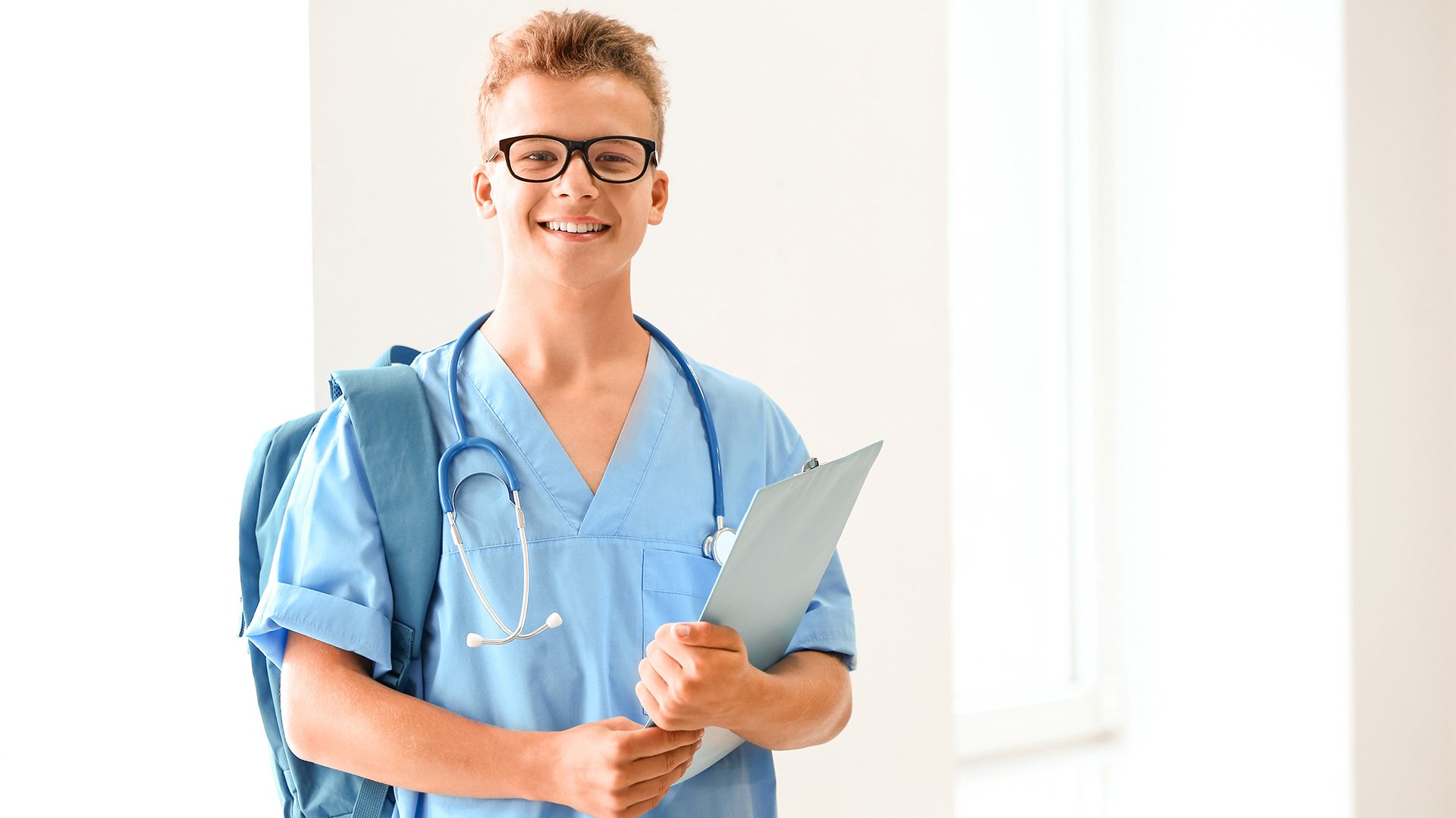 Medizinstudent im hellblauen Kasak trägt Rucksack Stethoskop und Klemmbrett.