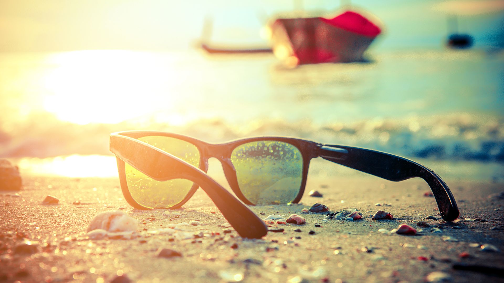 Sonnenbrille liegt im Sand am Strand. Auf dem Meer spiegelt sich die Sonne und im Hintergrund ist ein rotes Boot zu sehen.