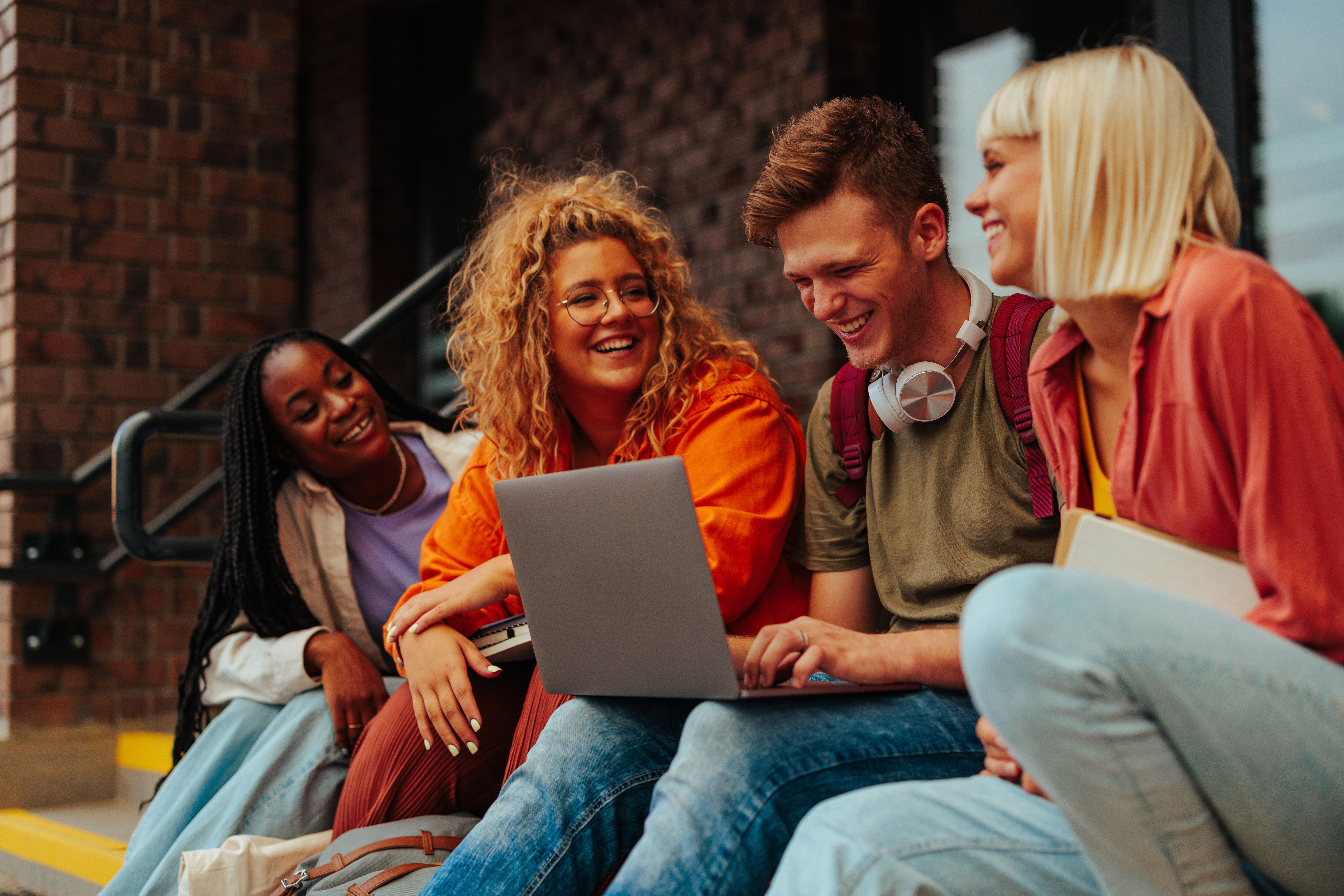 Studierende mit Laptop sitzen auf Treppe und lachen.  