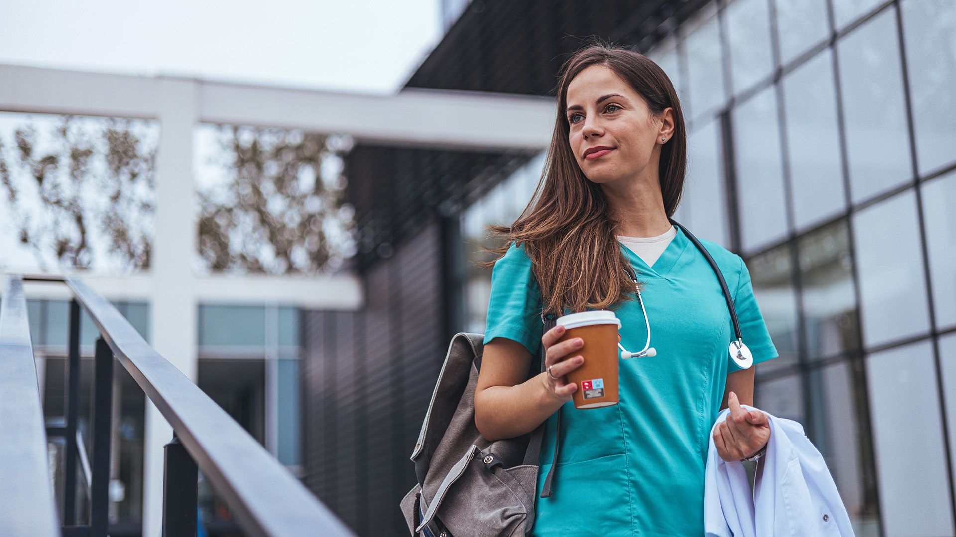 Ärztin vor dem Krankenhaus hält Kaffebecher und Kittel in der Hand und schaut zur Seite.