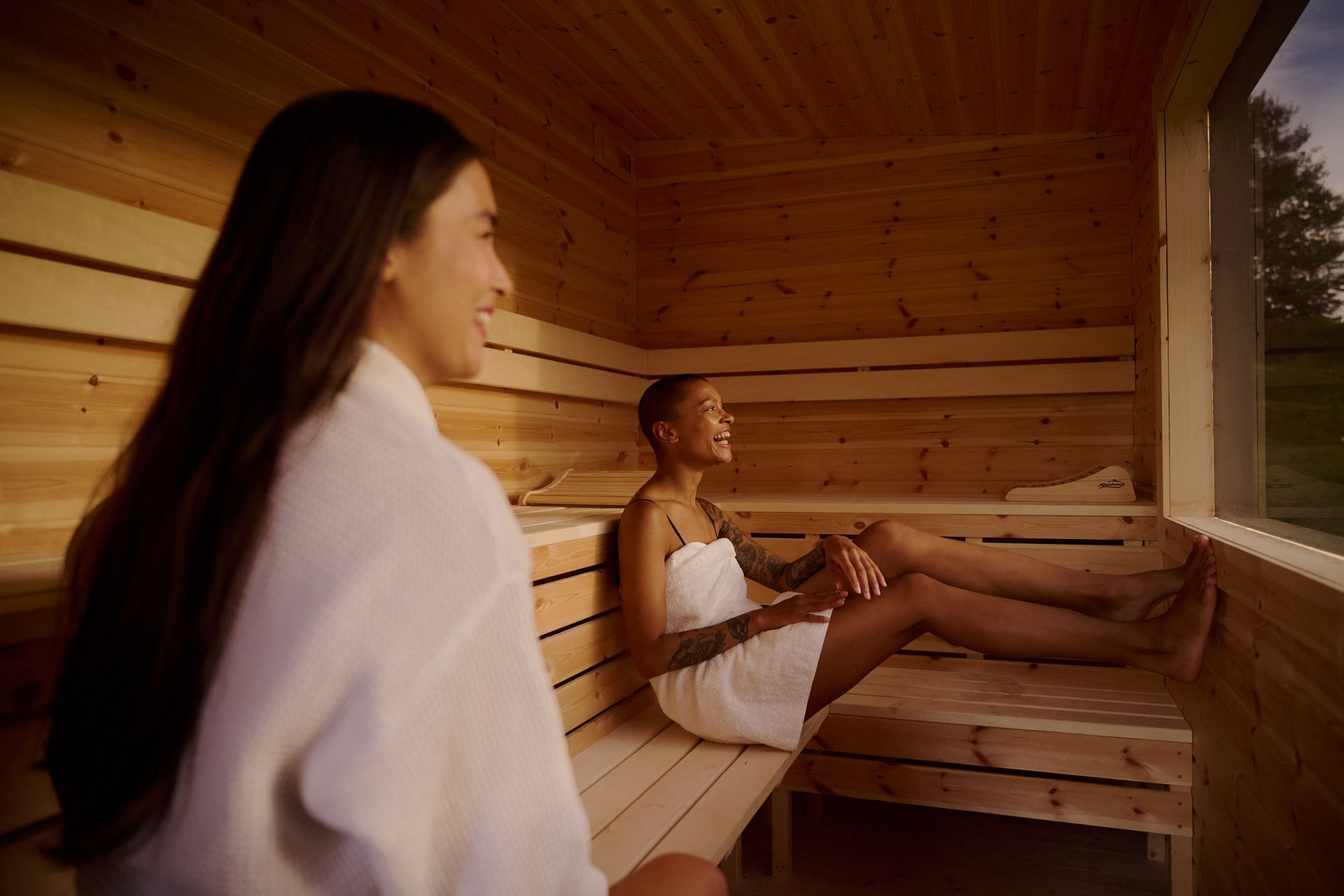 Two women, wearing bathrobes, sit happily in a sauna and relax.