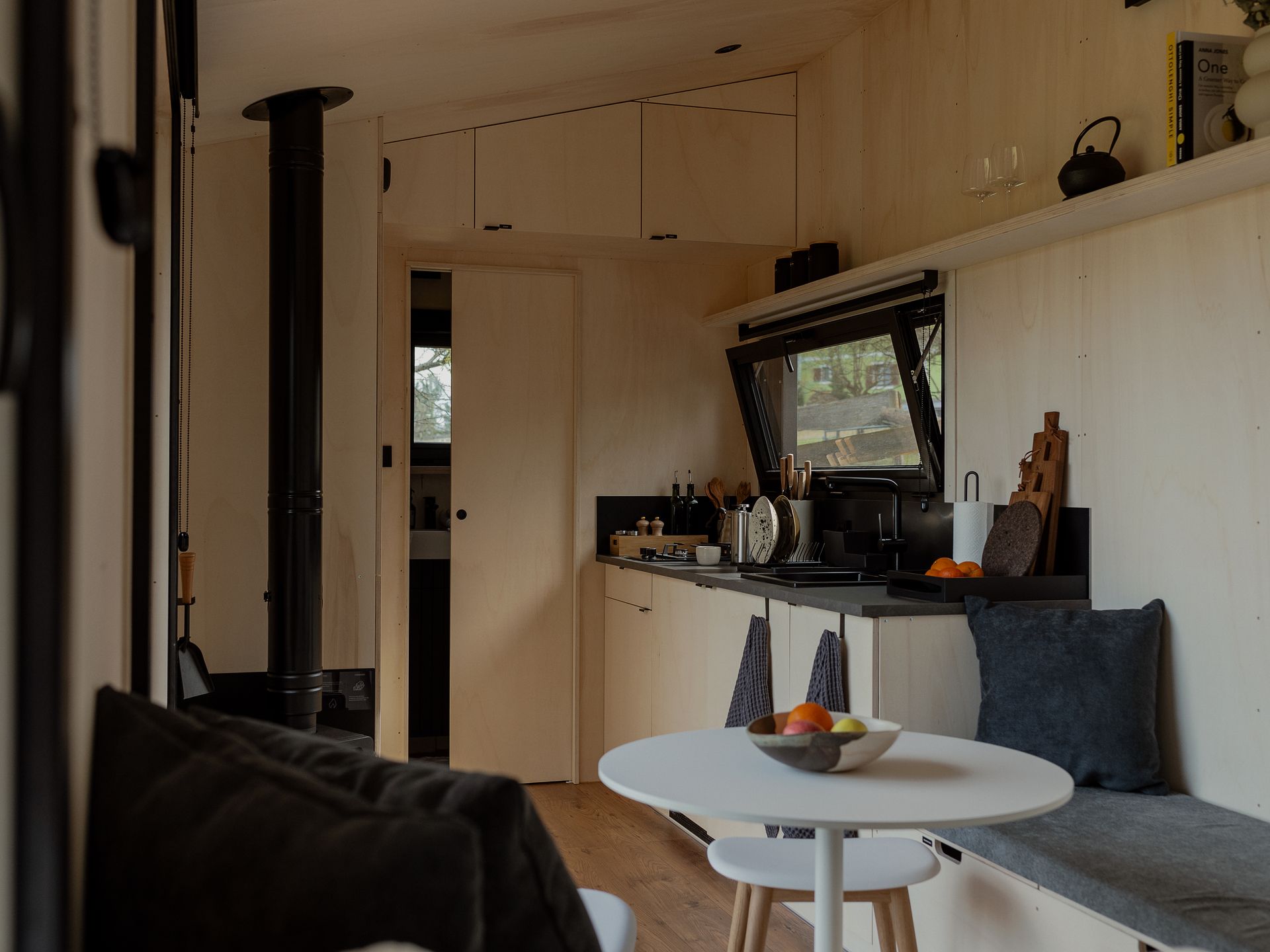 Blick vom Bett auf einen kleinen weißen Tisch mit einer Obstschale und die dahinter liegende Küchenzeile aus Holz