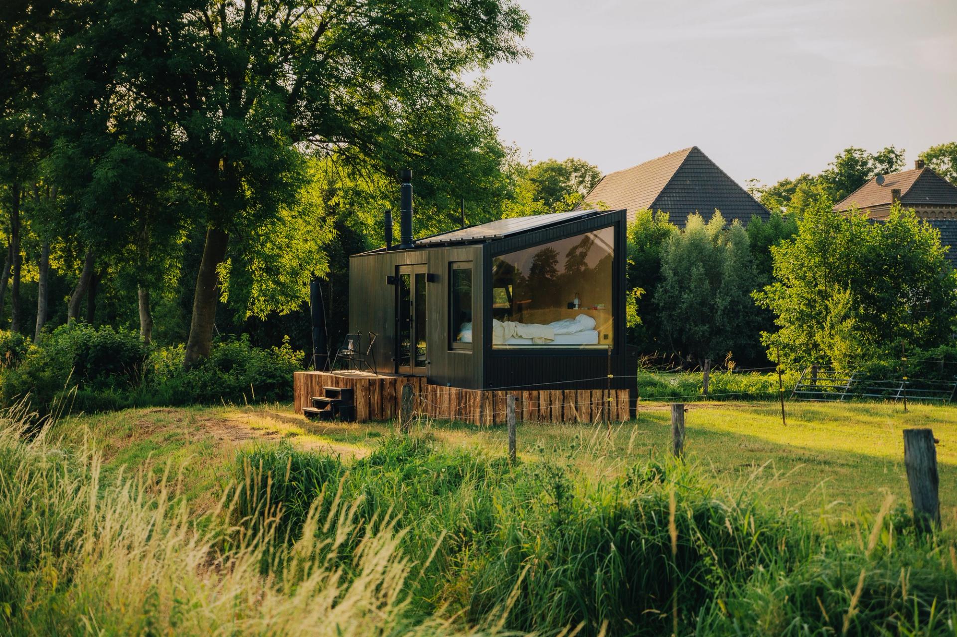 Black Raus glamping cabin in Germany with a large panoramic window, situated in a meadow surrounded by trees, with a farm visible in the background.