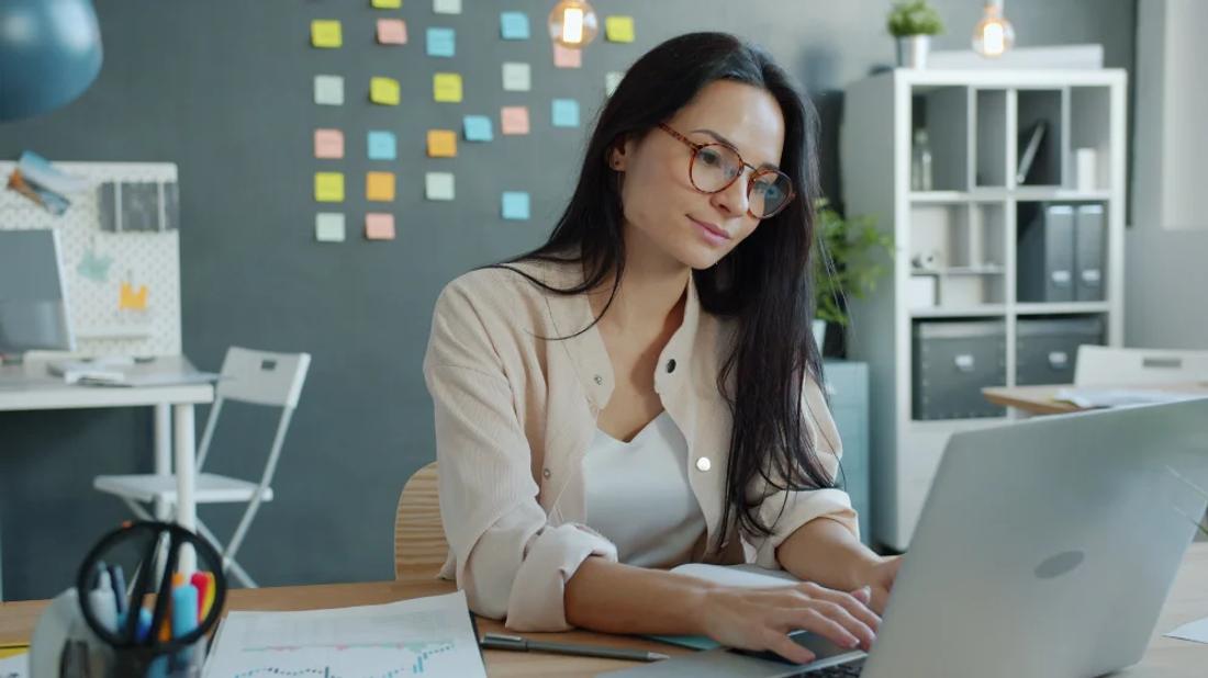 A woman in glasses smiling as she works on a laptop at her desk in a bright office