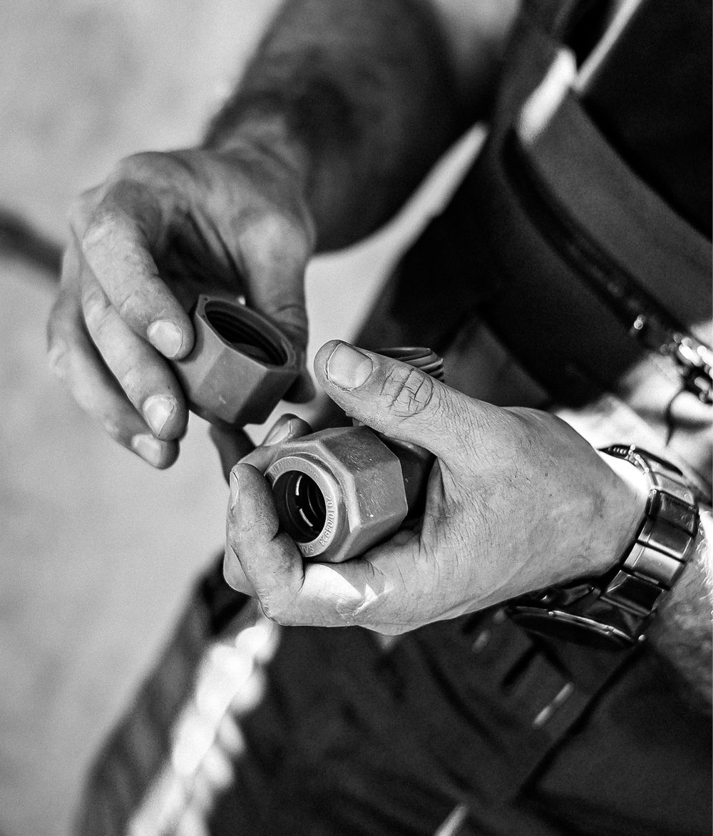 Black-and-white close-up of a plumber’s hands holding several Unitwist plumbing fittings, with a tool belt and workwear visible in the background.