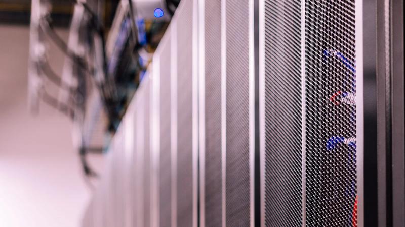 Filas de racks de servidores negros en un centro de datos, con fibra tendida entre los armarios.