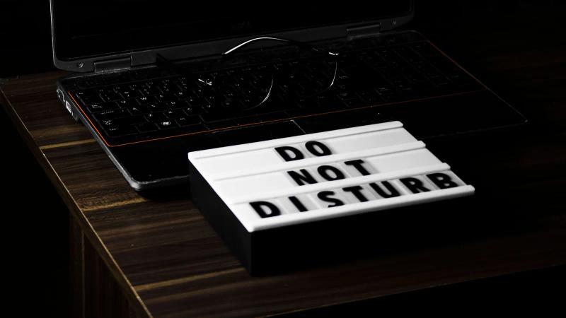 Un portátil sobre un escritorio en penumbra muestra la hora en pantalla, junto a un cartel de no molestar.
