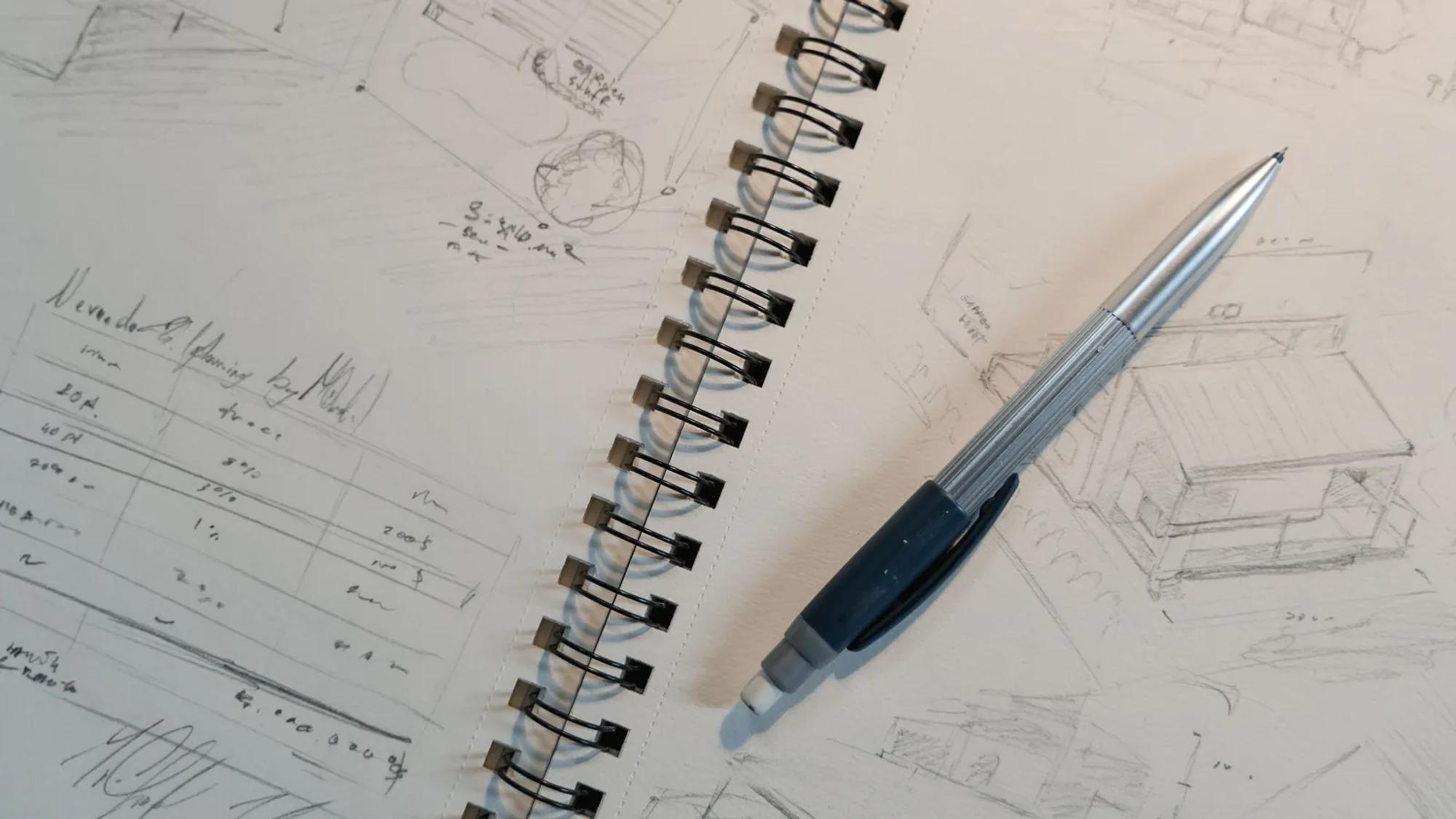 A mechanical pencil resting on a spiral notepad covered in small structural sketches drawn at thumbnail size.