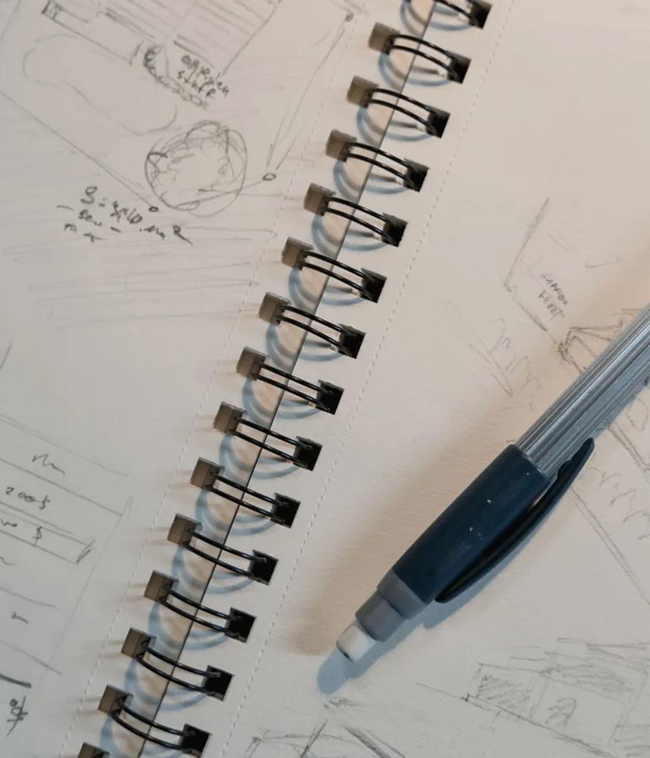 A mechanical pencil resting on a spiral notepad covered in small structural sketches drawn at thumbnail size.
