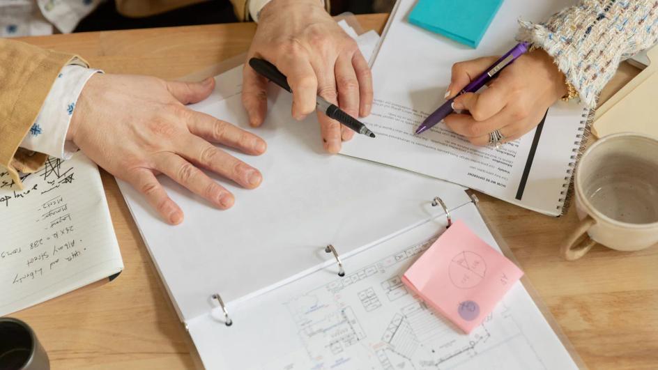 Two colleagues at a desk reading printed documents and marking them up during a project review