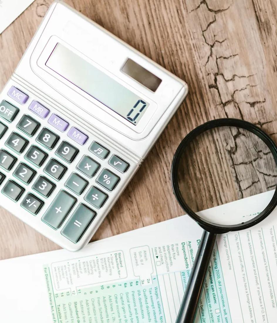Official forms laid out on a wooden desk with a calculator and a magnifying glass resting on the printed figures