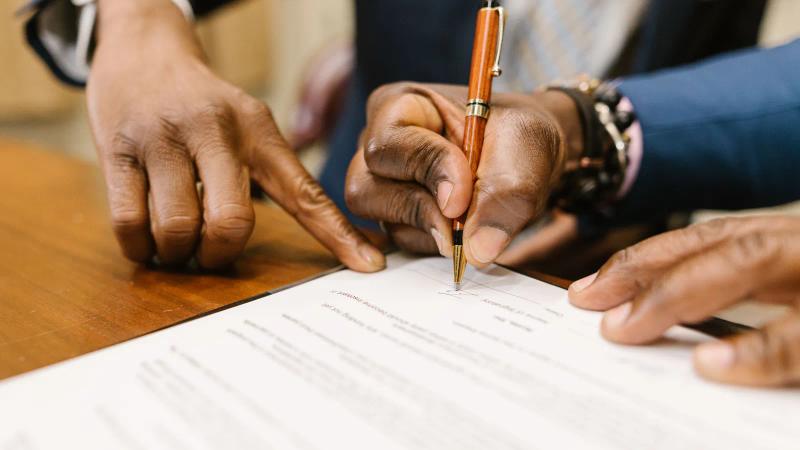 Una mano escribe sobre un contrato impreso en primer plano, con las condiciones del acuerdo bajo el bolígrafo.