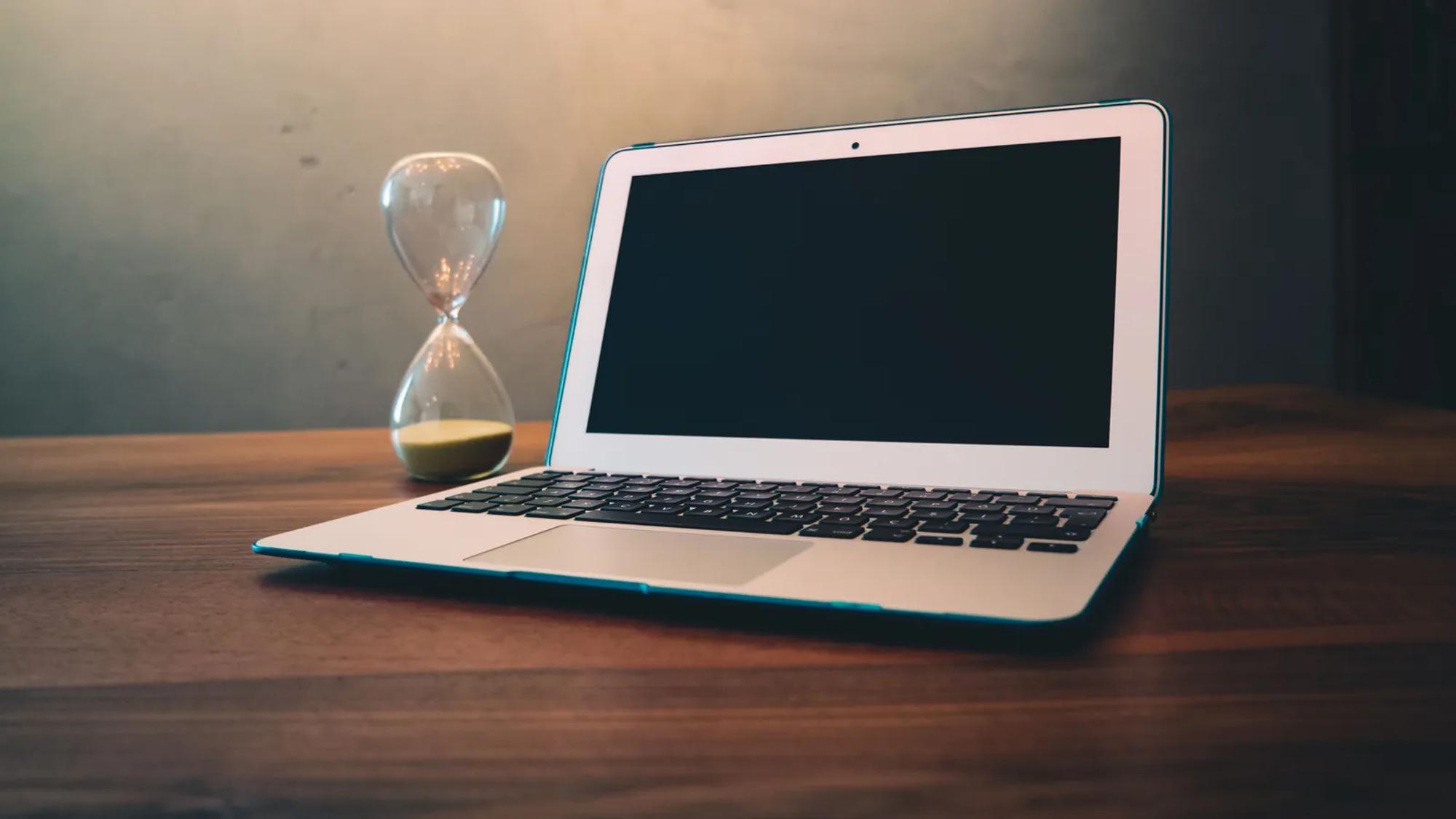 A laptop on a wooden desk beside an hourglass, the response clock running while nobody has been named as owner