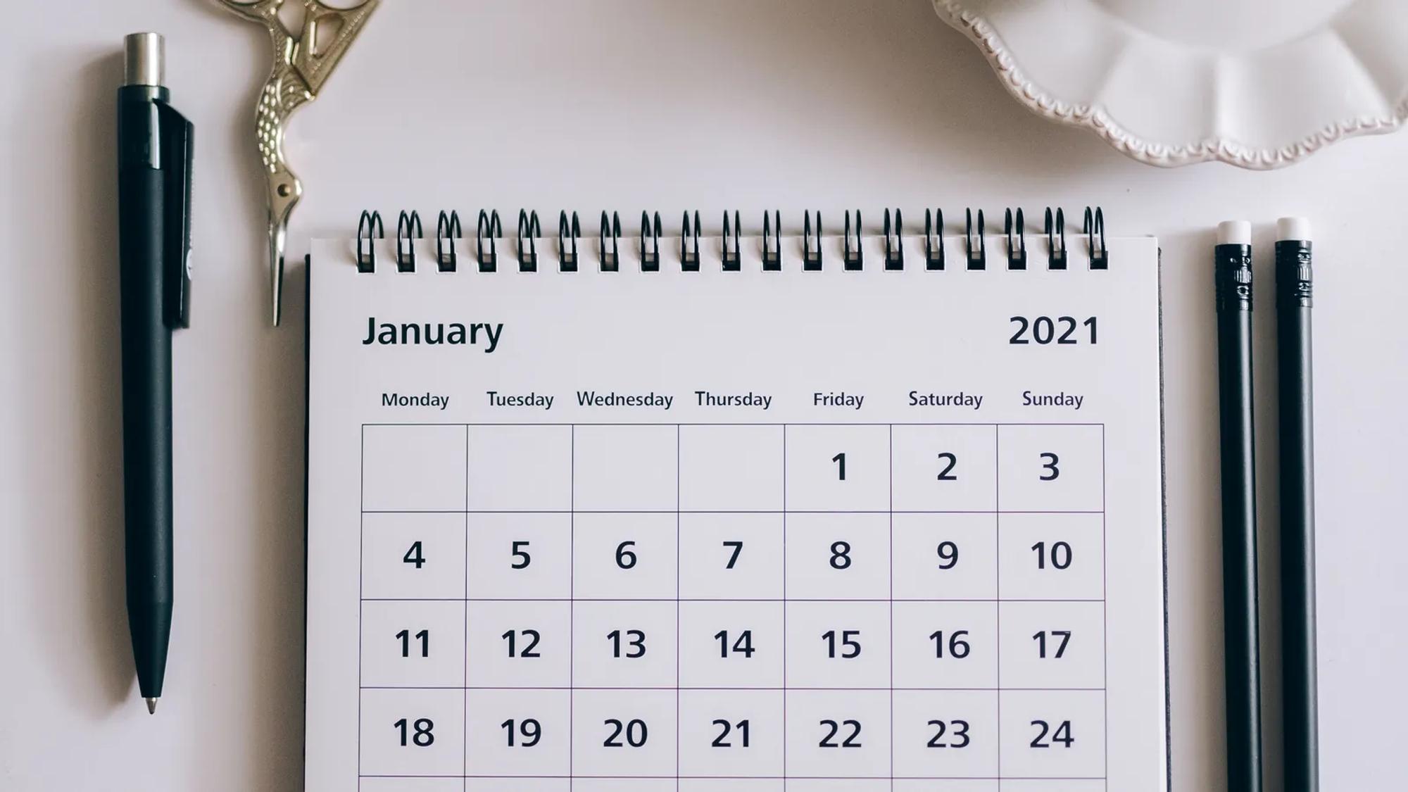 Calendario de mesa junto a un bolígrafo, clips y tijeras sobre una superficie blanca, listo para marcar fechas
