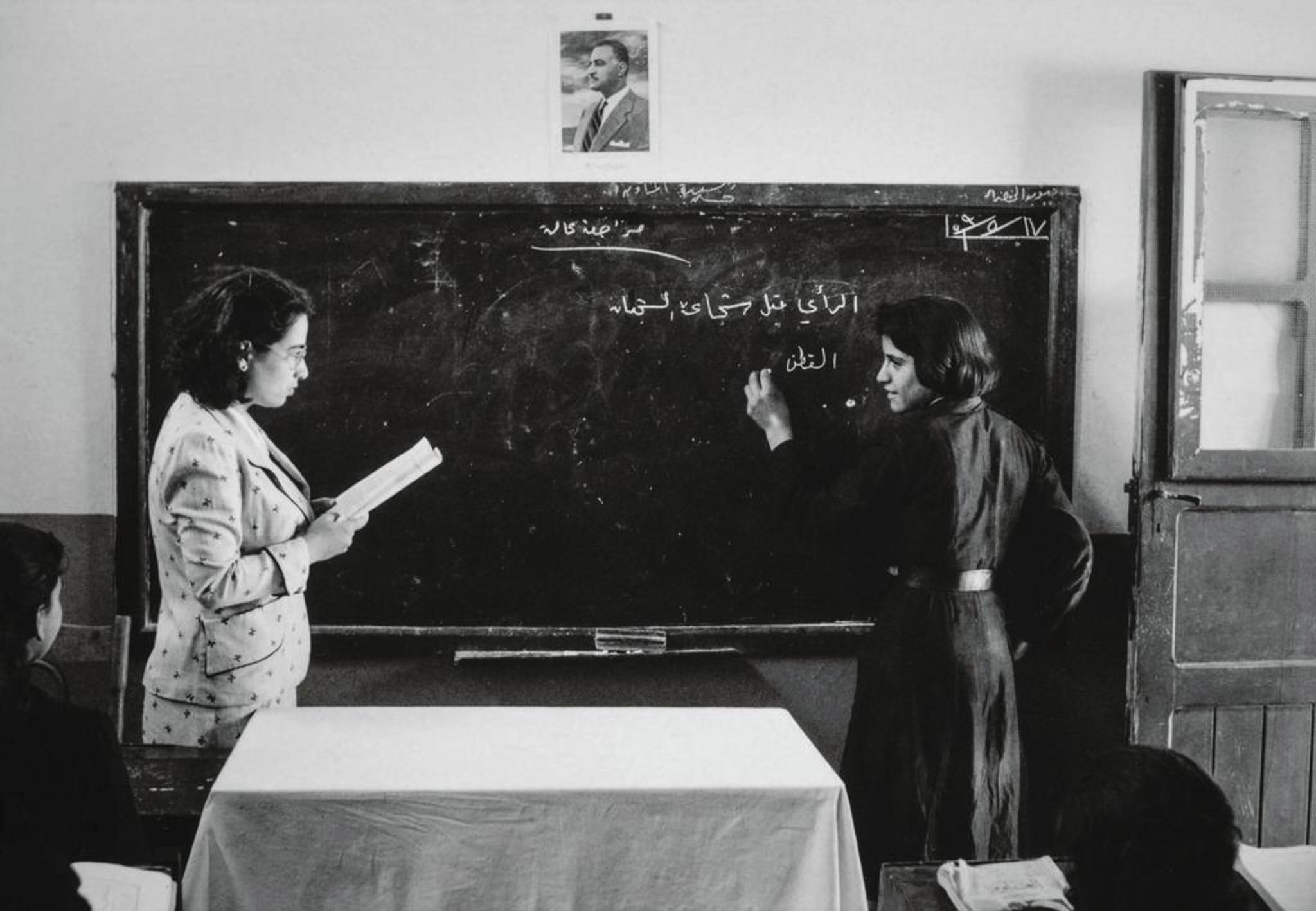 A female teacher and female student standing in front of a blackboard, they look at each other. The teacher holds a sheet of paper, the student writes on the board.