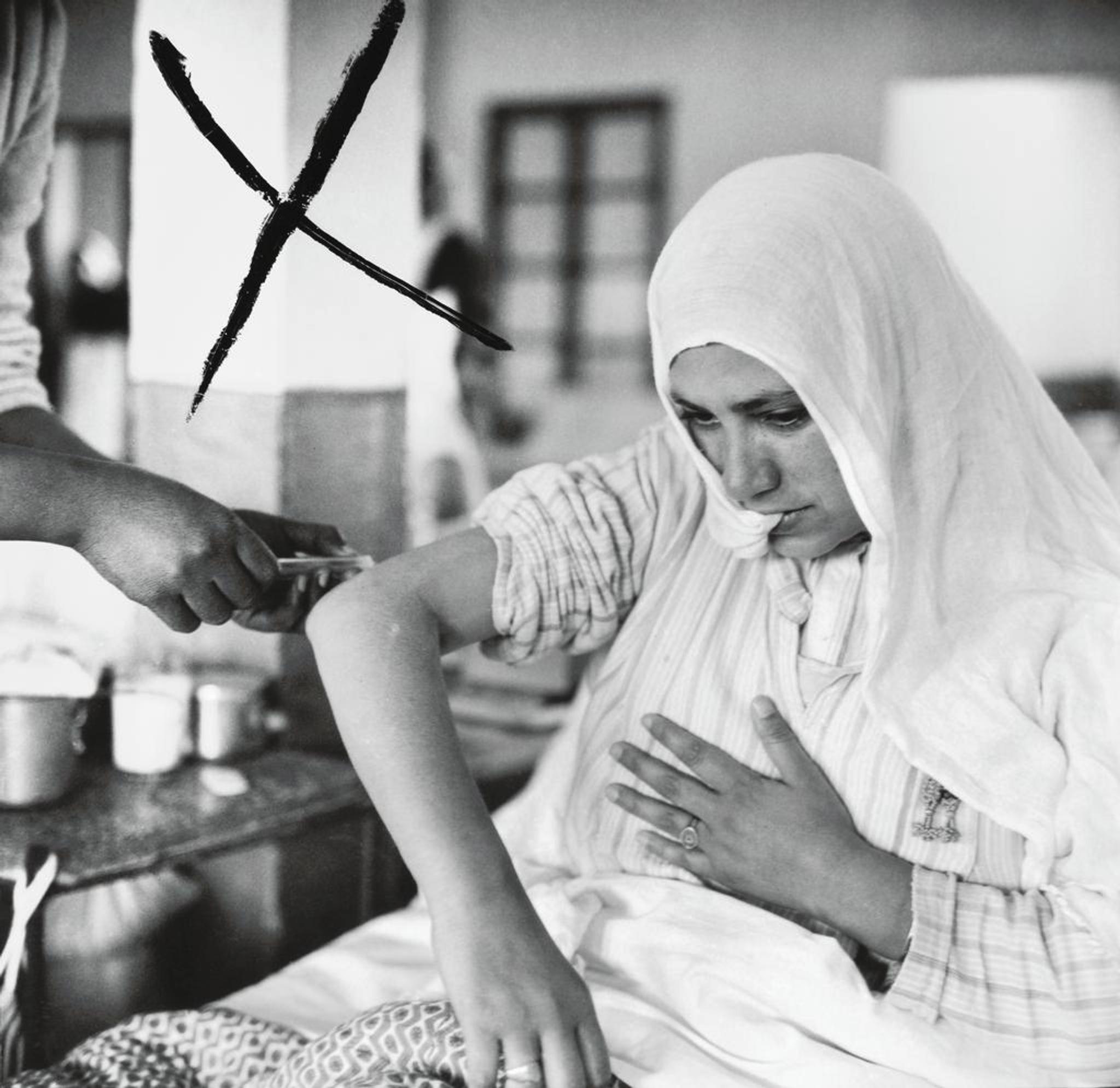 A woman with a shawl over her head gets an injection. A cross to the left of the image marks that the photographer has selected the image from a contact copy