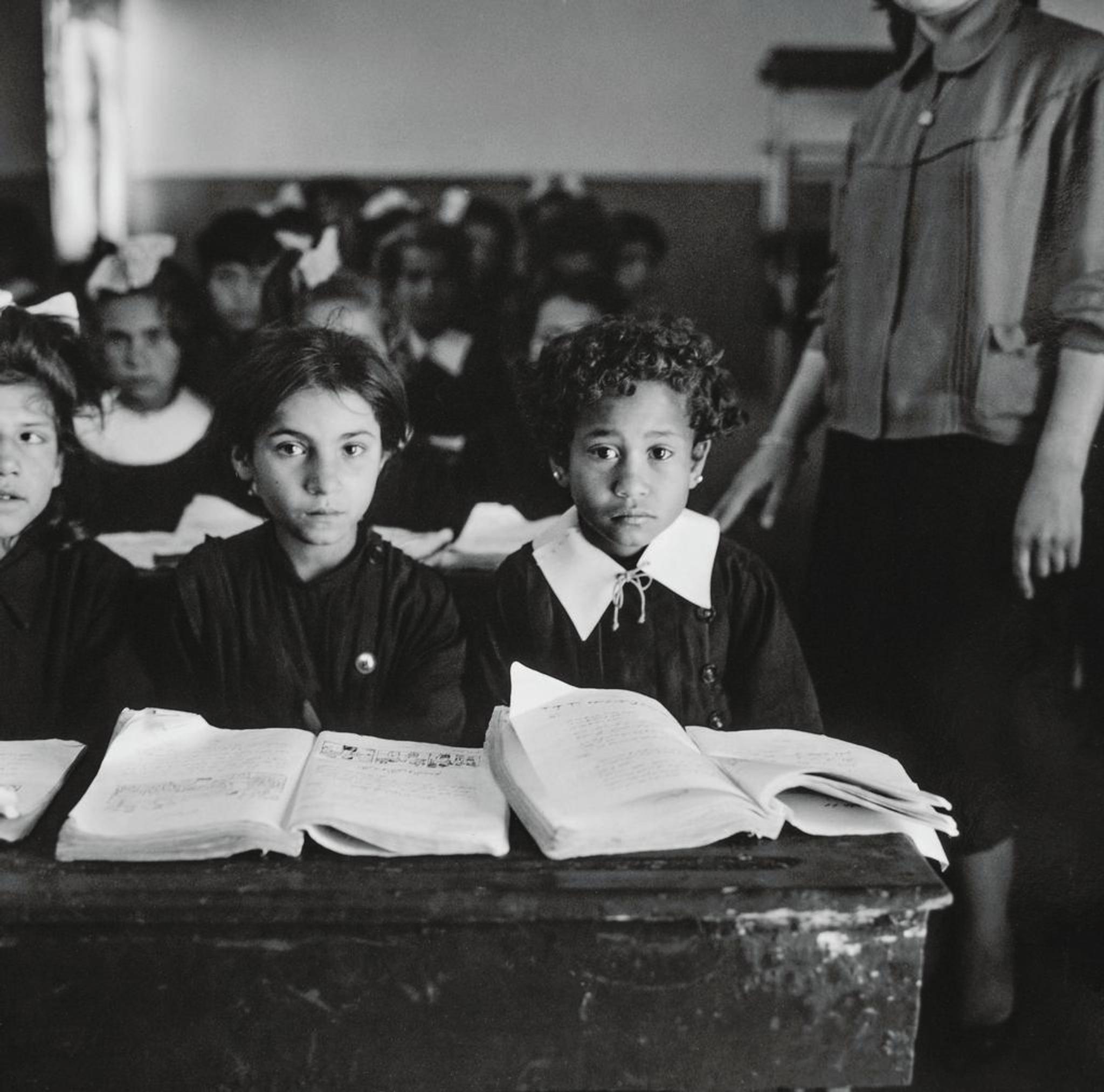A group of girls in a classroom, looking in the camera with serious faces. On the desks in front of them are open books.