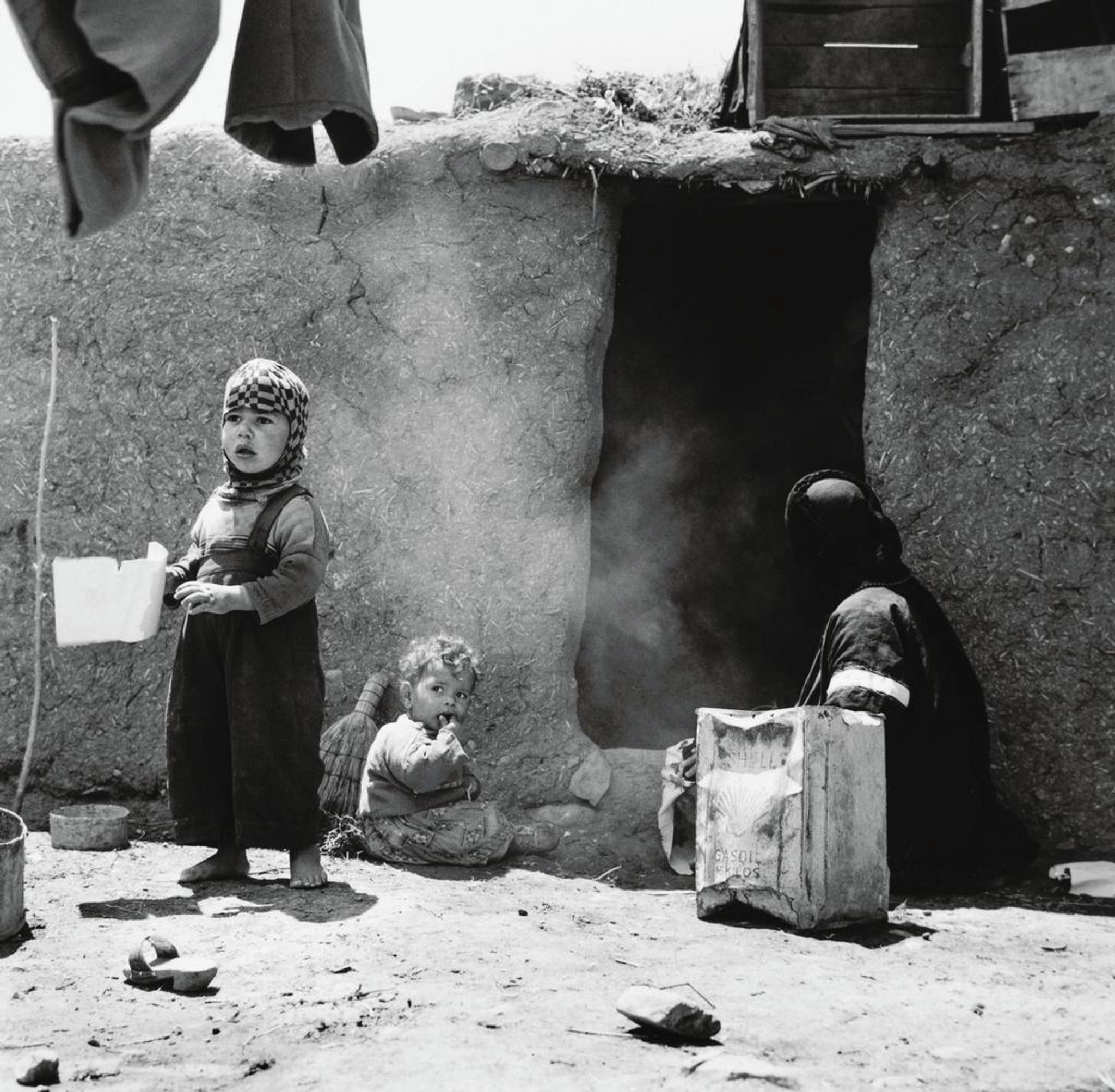 A women and two children in front of a clay house. The children are looking past the camera, the woman is facing the doorway.