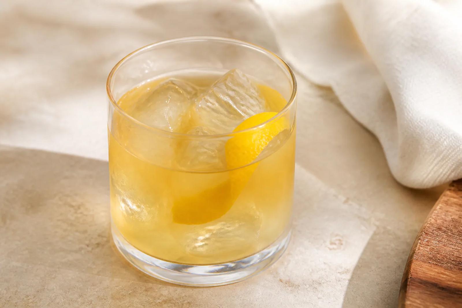 Pale gold Whiskey Sour in a rocks glass over fresh clear ice with a lemon peel and no foam.
