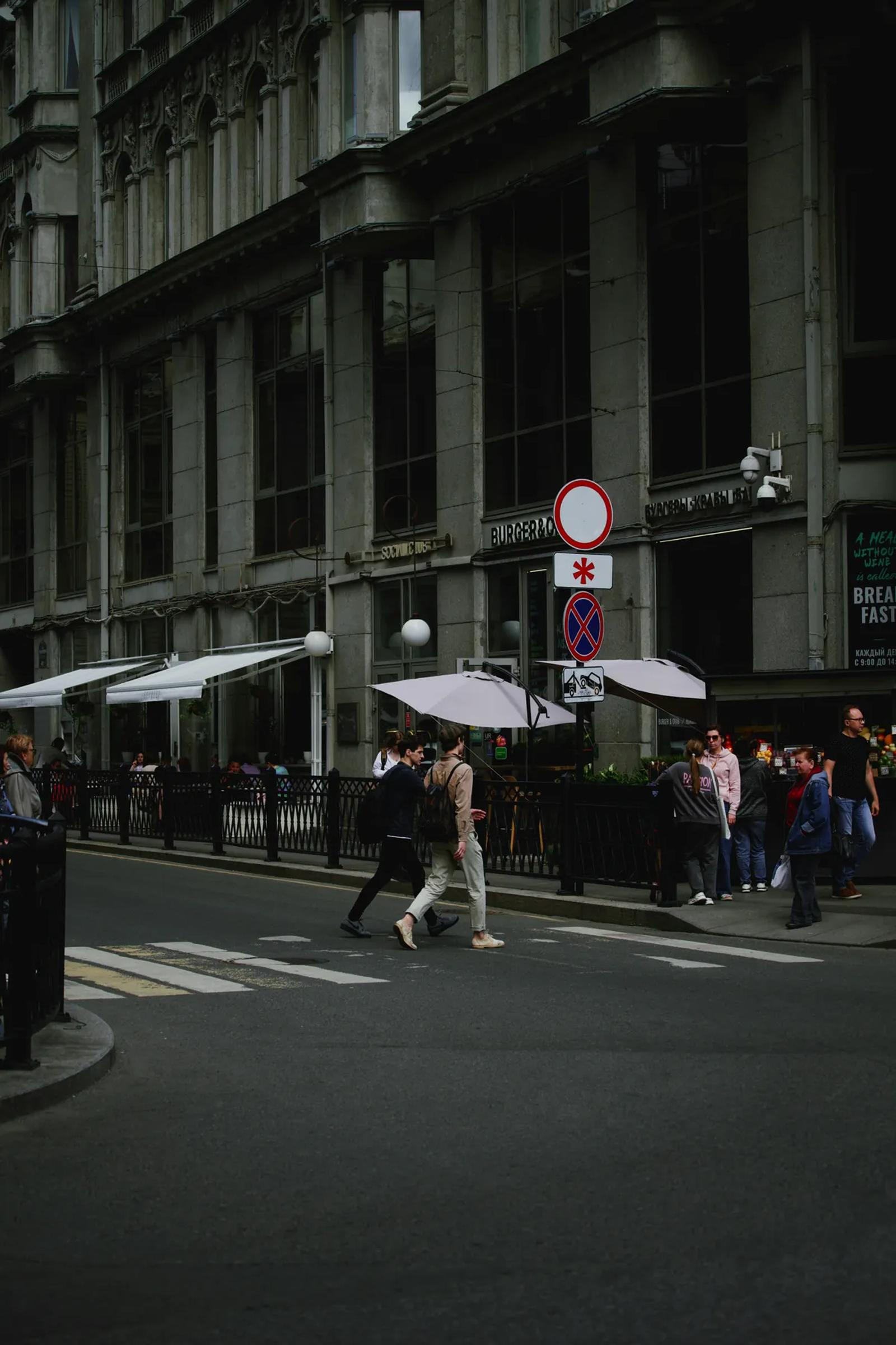 People walking along a shaded city sidewalk beside neighborhood storefronts