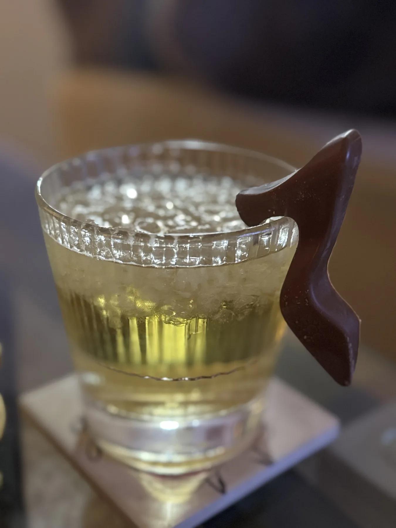 A pale cocktail in a textured rocks glass with a dark chocolate garnish