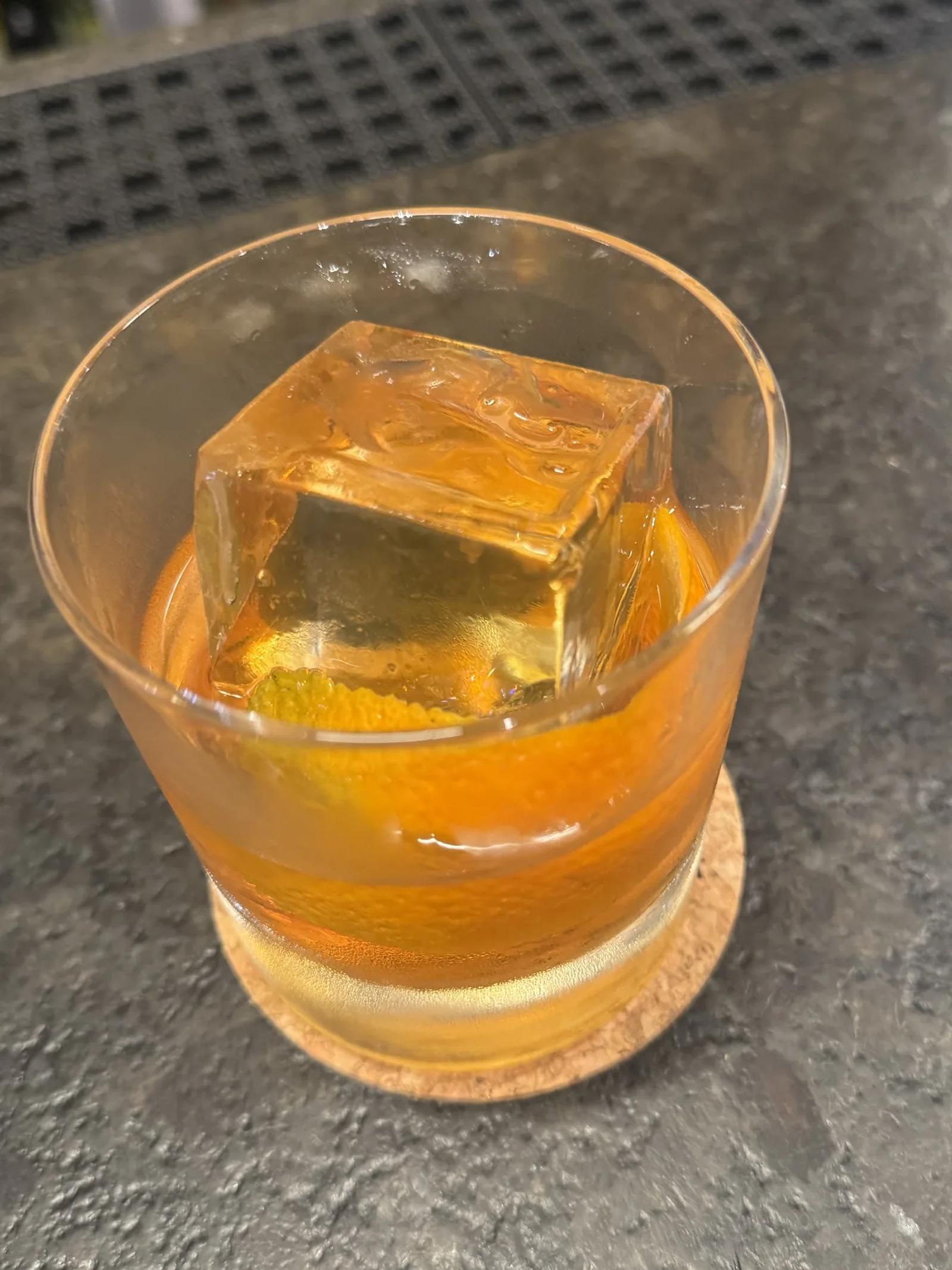 An amber drink with a large clear ice cube and citrus peel in a rocks glass
