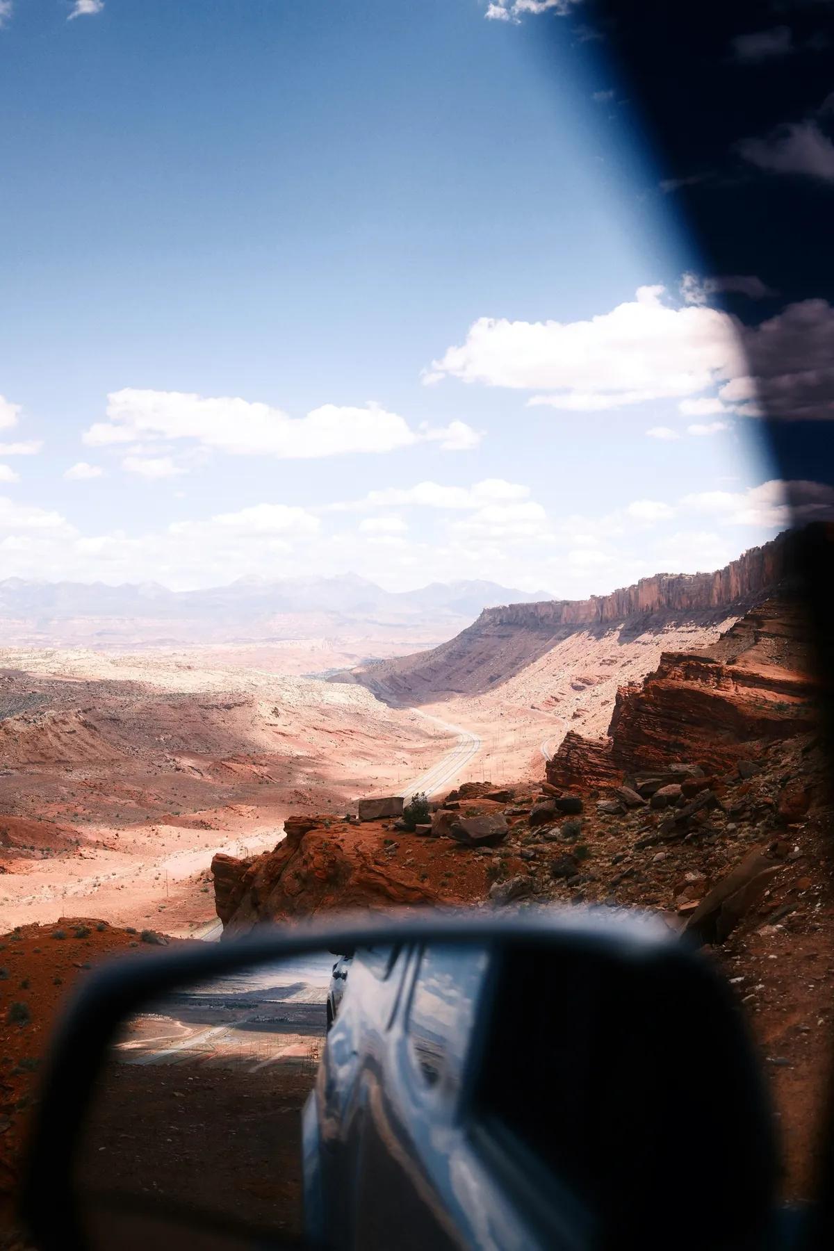 A white SUV following a two-lane road between sunlit red-rock walls near Moab, Utah